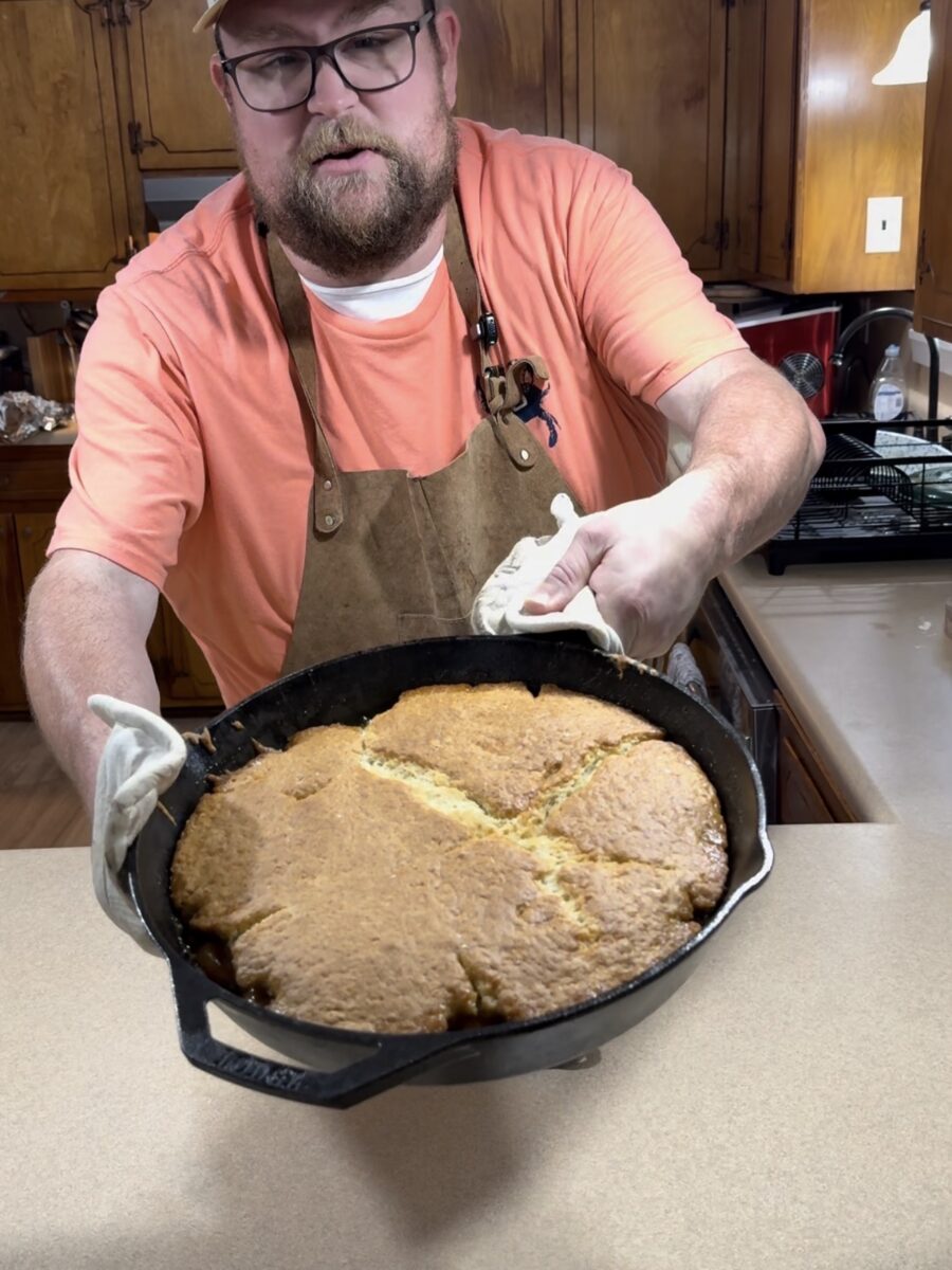 A man wearing glasses, a cap, and an apron holds out a cast iron skillet with freshly baked cornbread in a kitchen.