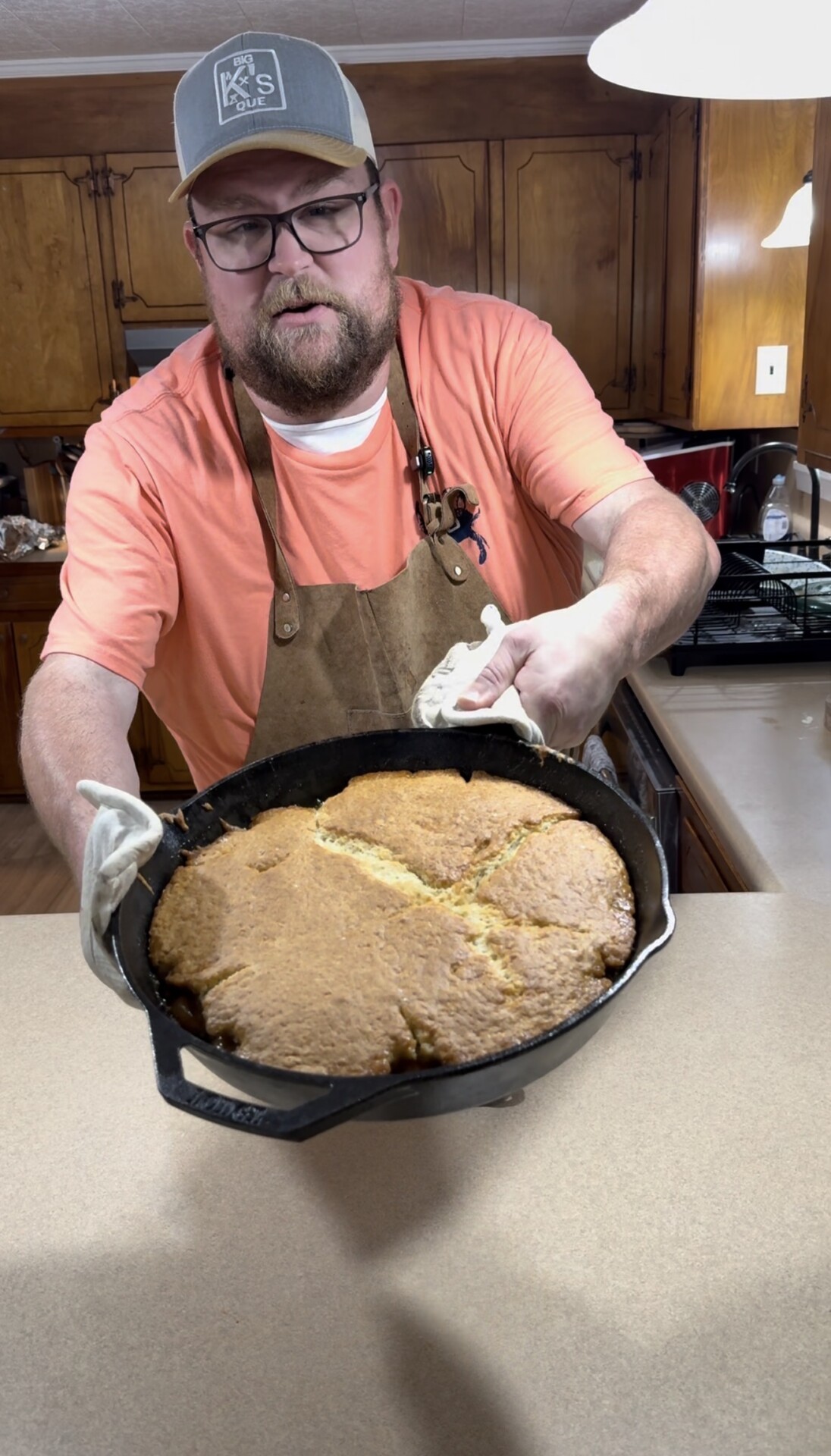 A man in an apron and cap holds a large skillet of freshly baked cornbread in a kitchen, presenting it toward the camera with a slight smile.