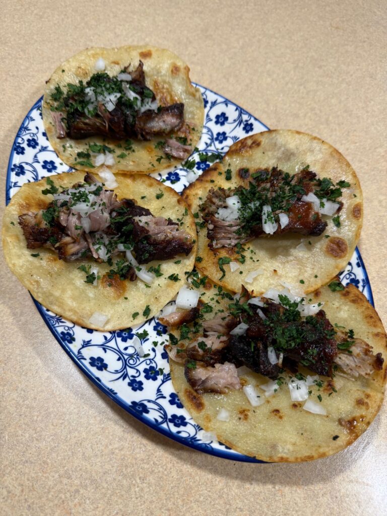 Four tacos with chopped meat, cilantro, and diced onions on corn tortillas are arranged on a blue and white patterned plate, set on a light brown countertop.