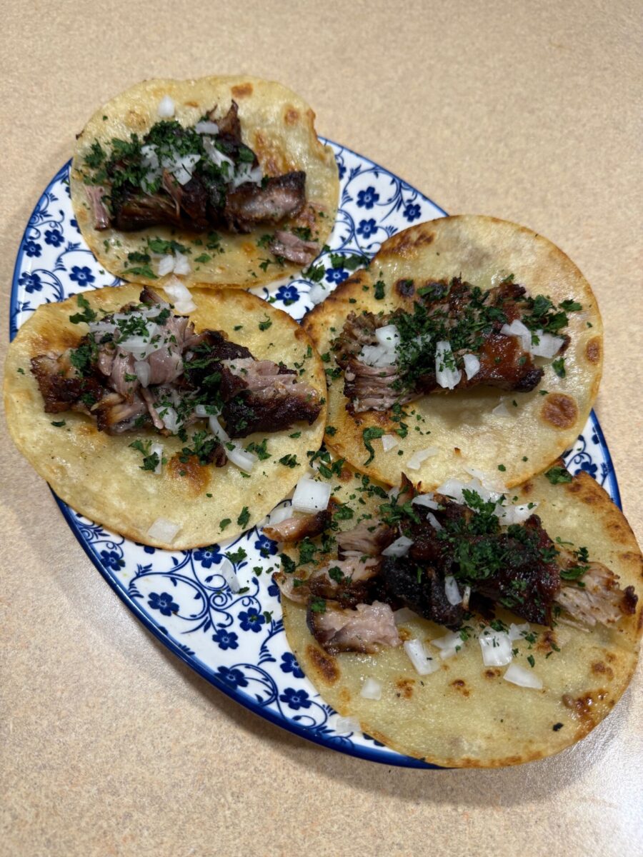 Four tacos with chopped meat, cilantro, and diced onions on corn tortillas are arranged on a blue and white patterned plate, set on a light brown countertop.