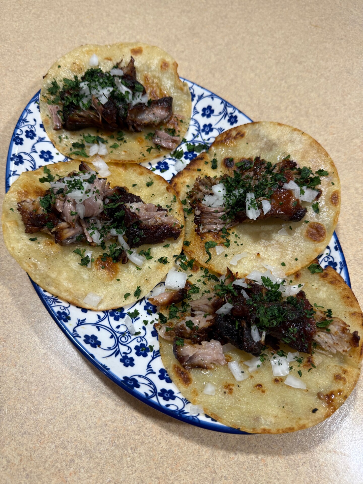 Four tacos with chopped meat, cilantro, and diced onions on corn tortillas are arranged on a blue and white patterned plate, set on a light brown countertop.