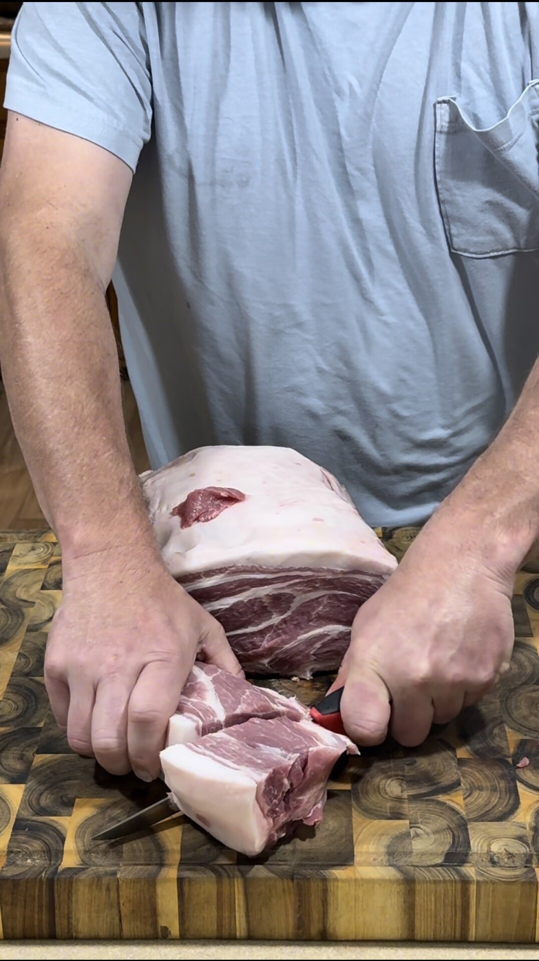 A person wearing a light blue shirt uses a knife to cut a large piece of raw pork on a patterned wooden cutting board.