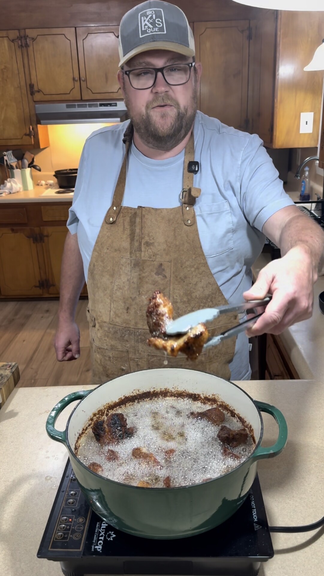 A man wearing a brown apron and a cap stands in a kitchen, holding a piece of fried food with tongs over a green pot of sizzling oil on the stove.