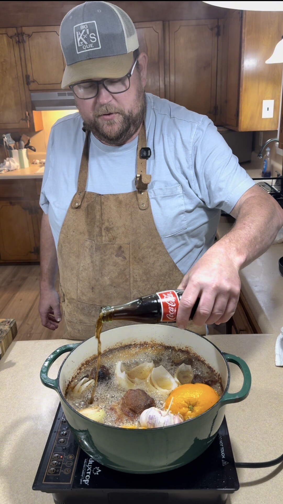 A man in a cap and apron pours Coca-Cola into a large green pot filled with meat, onions, garlic, and an orange, cooking on a stovetop in a rustic kitchen.