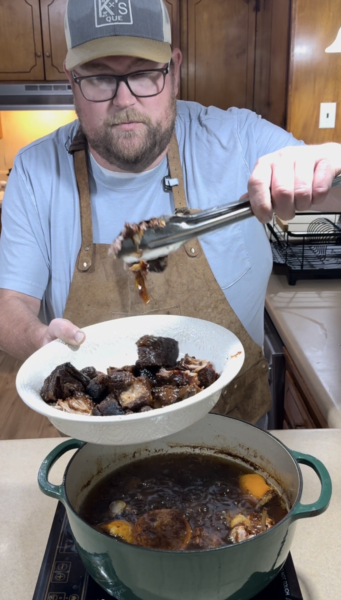 A man wearing glasses and a brown apron uses tongs to serve braised meat from a pot into a white bowl in a kitchen.