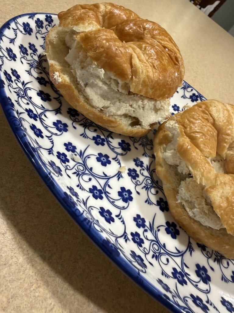 Two croissant sandwiches filled with a creamy chicken salad sit on a decorative white plate with blue floral patterns, placed on a tan countertop.