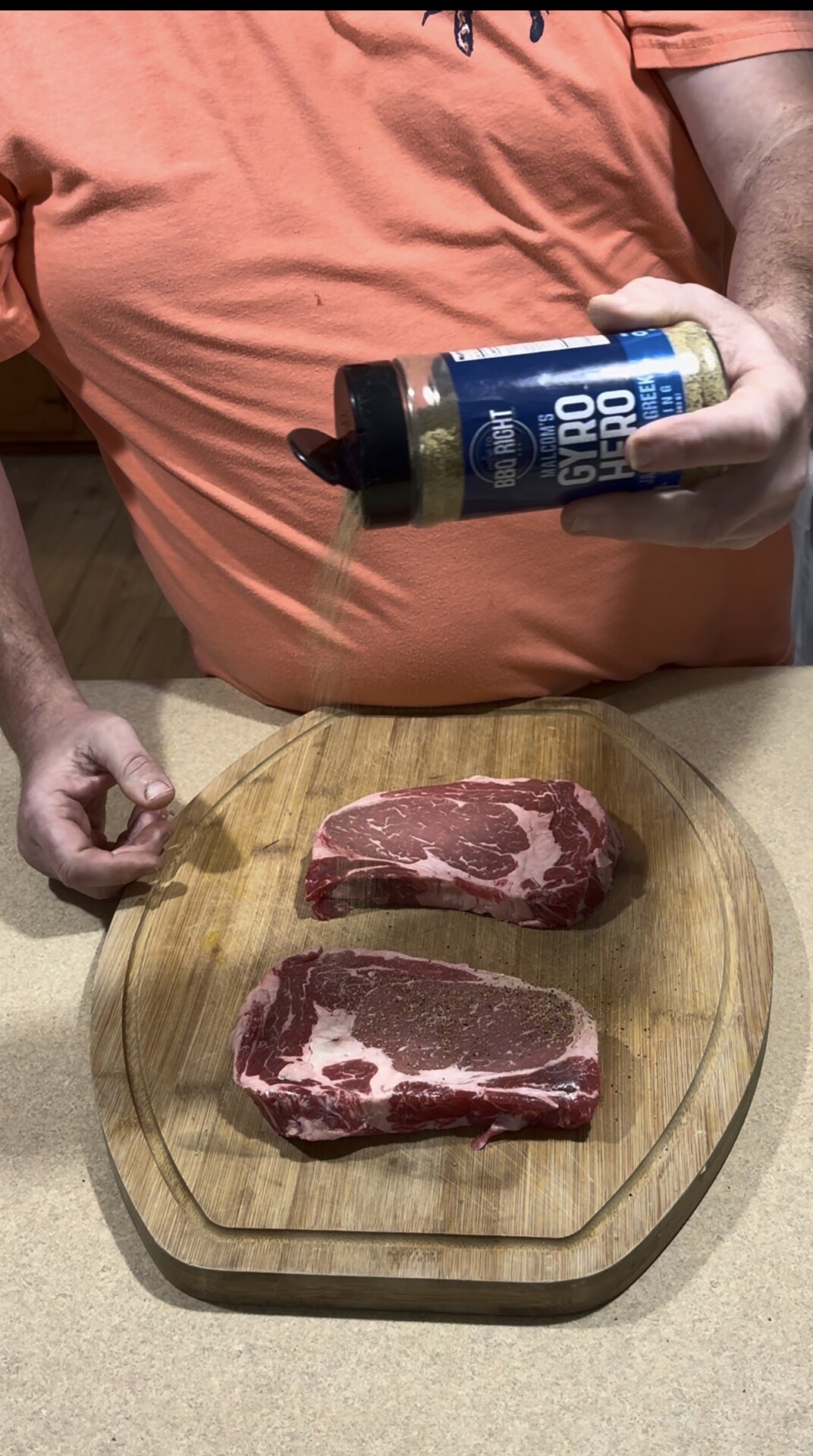 A person in an orange shirt sprinkles seasoning from a blue container onto two raw ribeye steaks placed on a wooden cutting board.