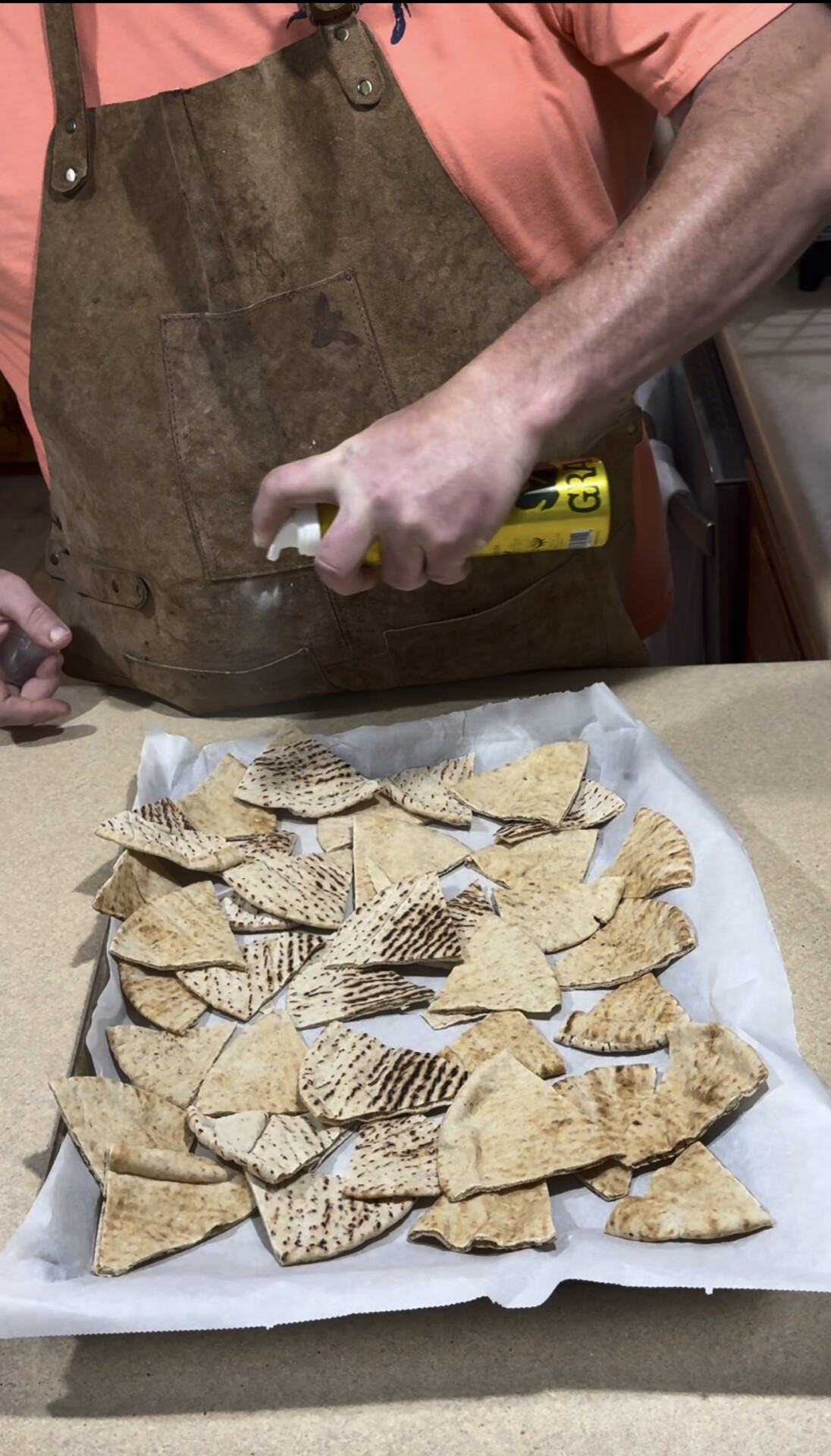 A person wearing a brown apron sprays cooking oil onto pieces of pita bread spread out on a parchment-lined baking tray on a kitchen counter.