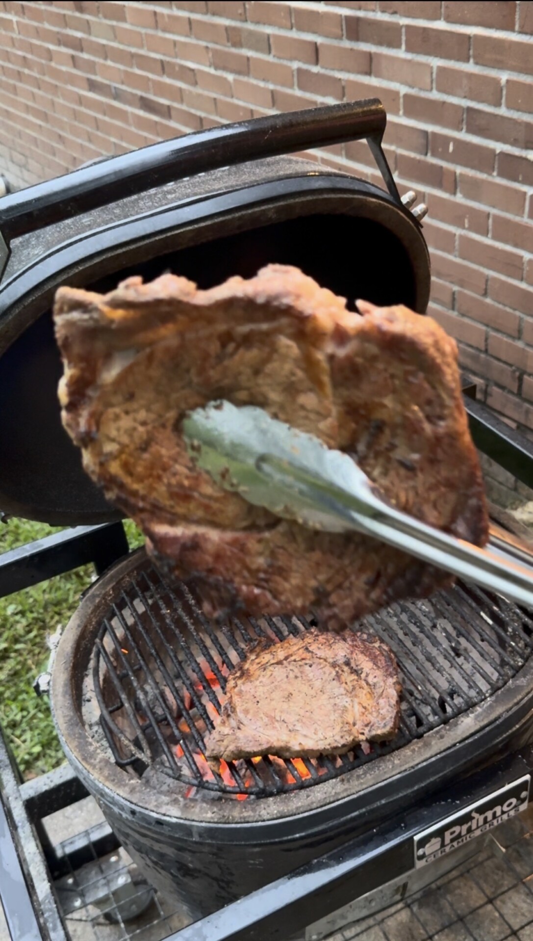 A close-up of tongs holding a grilled steak above a barbecue grill, with another steak cooking over glowing coals. The grill is outdoors near a brick wall.