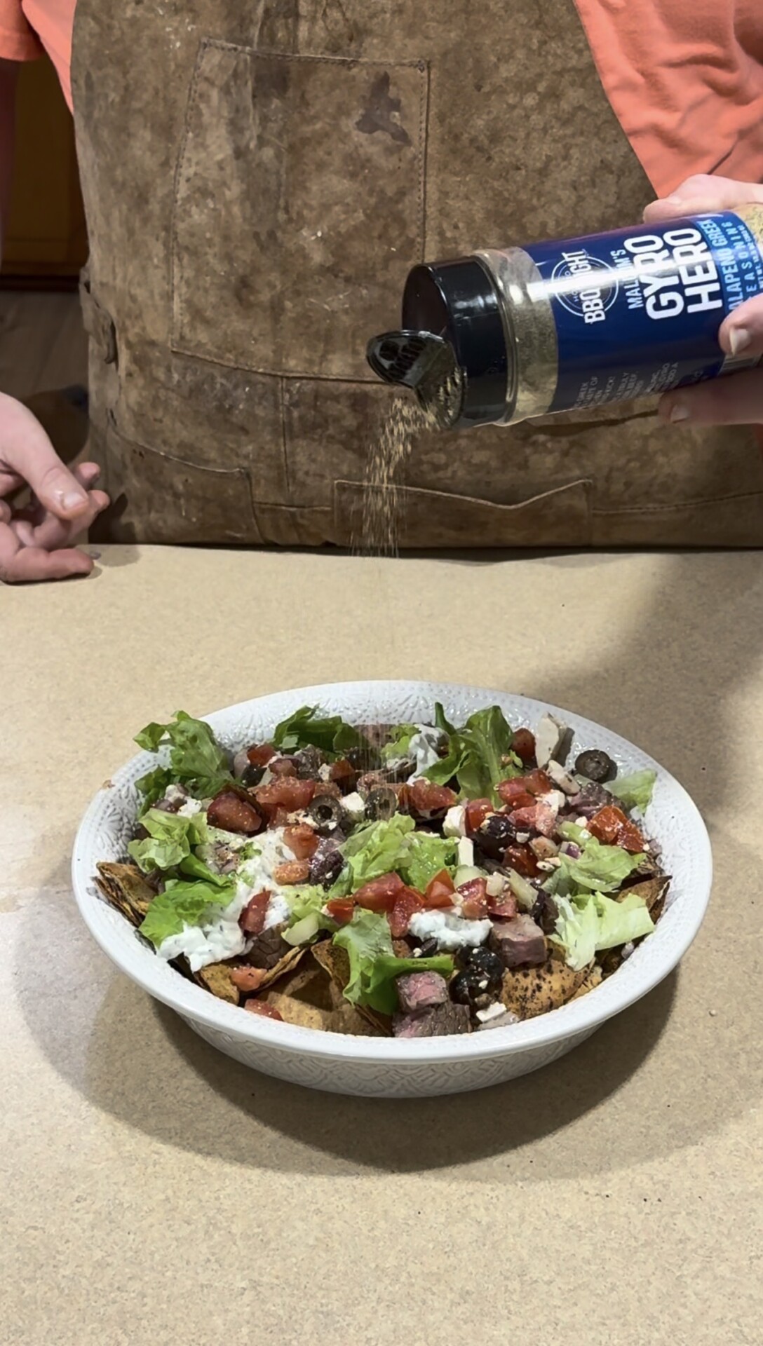 A person sprinkles seasoning from a container labeled Gyro Hero onto a bowl of salad with lettuce, tomatoes, meat, olives, and sauce on a countertop.