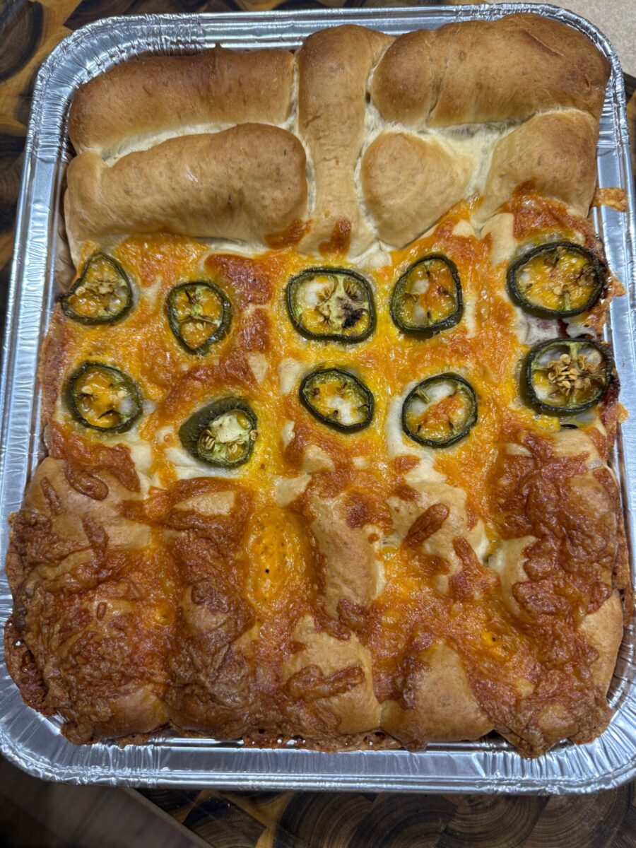 A baked focaccia bread in a rectangular foil tray, topped with melted cheddar cheese and sliced jalapeños. The bread is golden brown with a slightly puffy and airy texture.