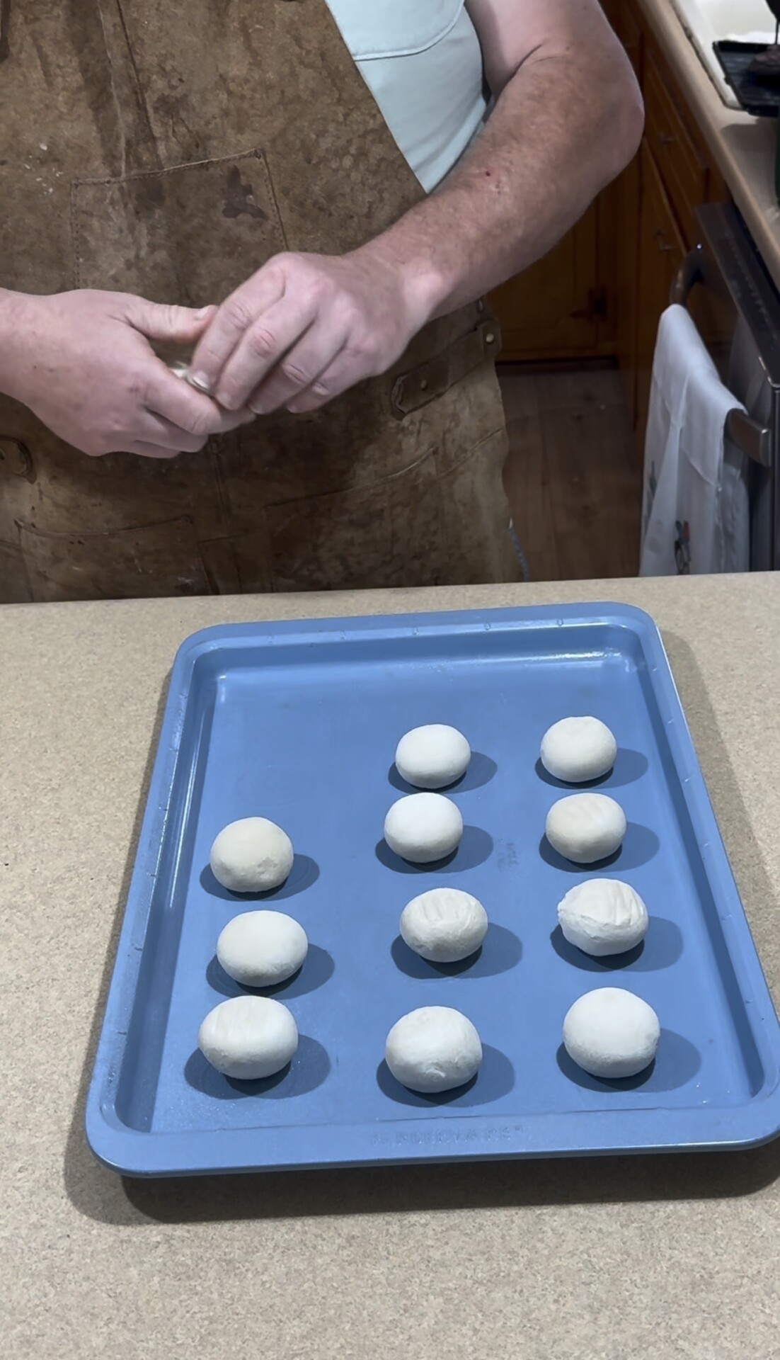 A person shapes dough balls by hand, with twelve evenly spaced dough balls on a blue baking tray placed on a kitchen counter.