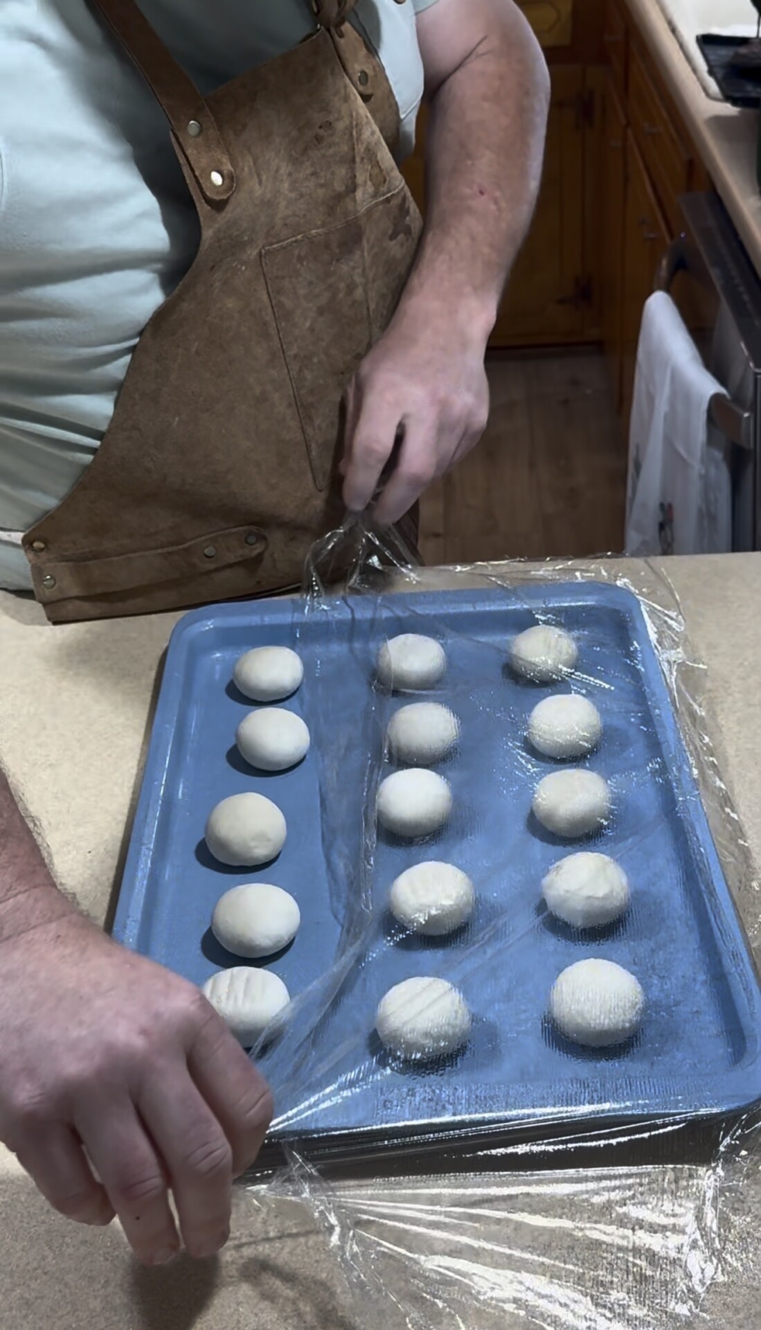 A person wearing a brown apron covers a blue baking sheet with plastic wrap. The baking sheet holds 15 evenly spaced dough balls on a kitchen counter.
