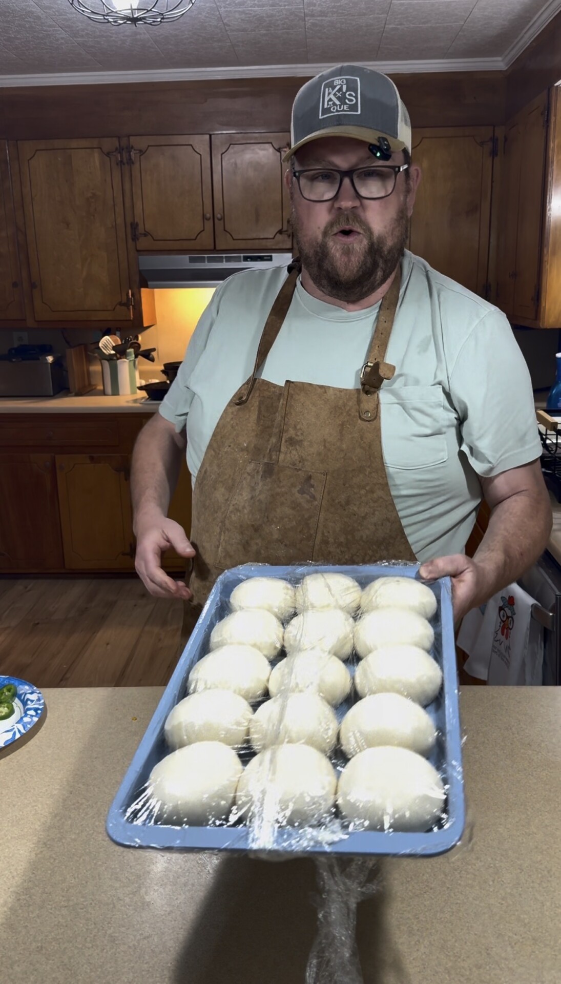A bearded man wearing glasses, a cap, and a brown apron holds a tray of dough rolls covered in plastic wrap in a kitchen with wooden cabinets.