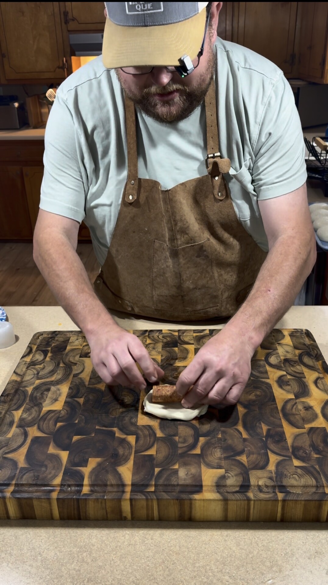 A person wearing a tan apron and light blue shirt is rolling a piece of food, possibly meat, on a cutting board with a swirled wood pattern in a kitchen setting.