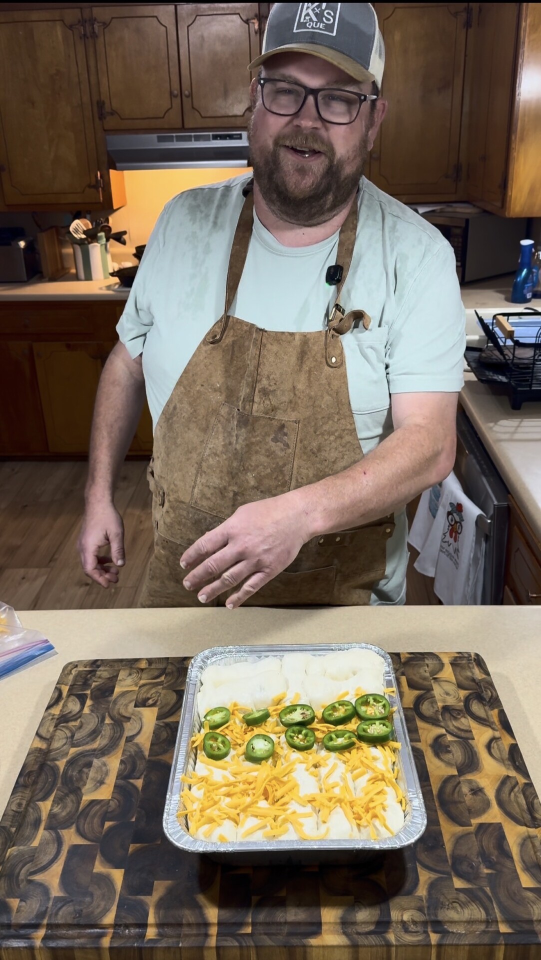 A man wearing a brown apron and cap stands in a kitchen, smiling at the camera. In front of him is a baking dish filled with shredded cheese, sliced onions, and jalapeños, placed on a wooden cutting board.