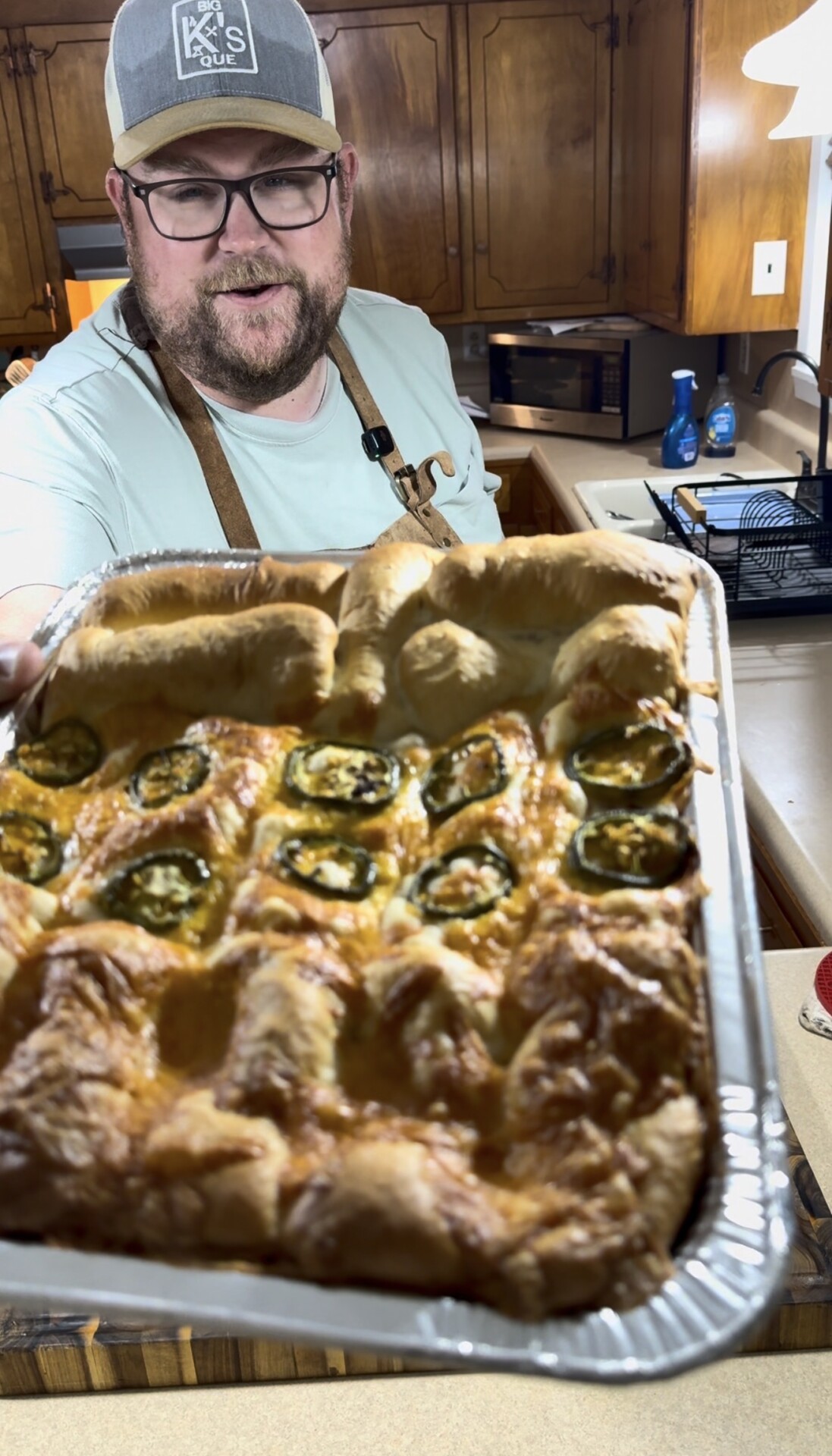 A bearded man wearing glasses, a hat, and a light blue shirt presents a tray of freshly baked, golden-brown bread topped with sliced jalapeños in a kitchen setting.