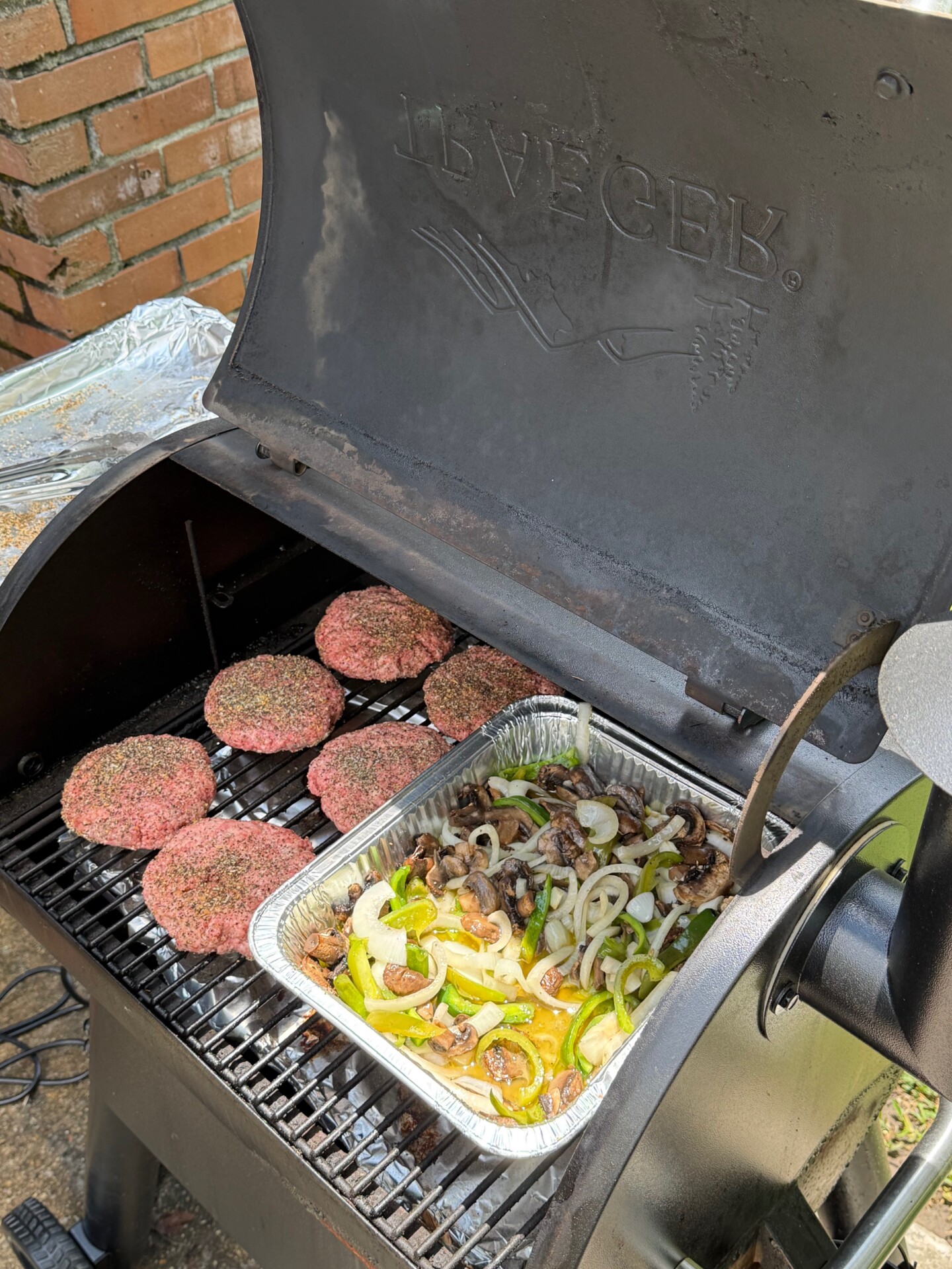 Six raw burger patties and a foil tray filled with sliced onions, green peppers, and mushrooms are cooking on a Traeger grill outside, with a brick wall visible in the background.