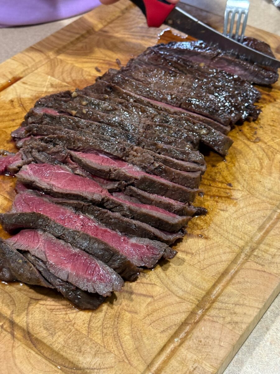 Sliced medium-rare steak on a wooden cutting board, with some pieces showing a pink center. A person is holding a fork and knife, about to cut another piece.