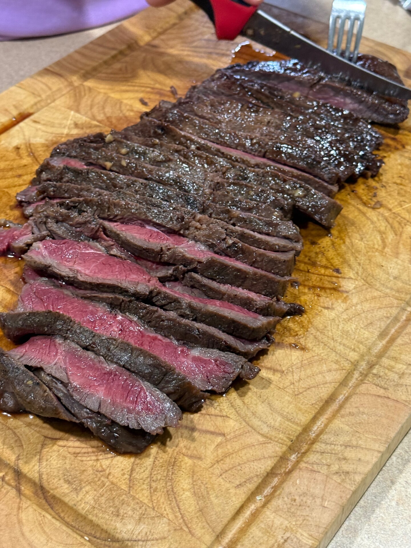 Sliced medium-rare steak on a wooden cutting board, with some pieces showing a pink center. A person is holding a fork and knife, about to cut another piece.