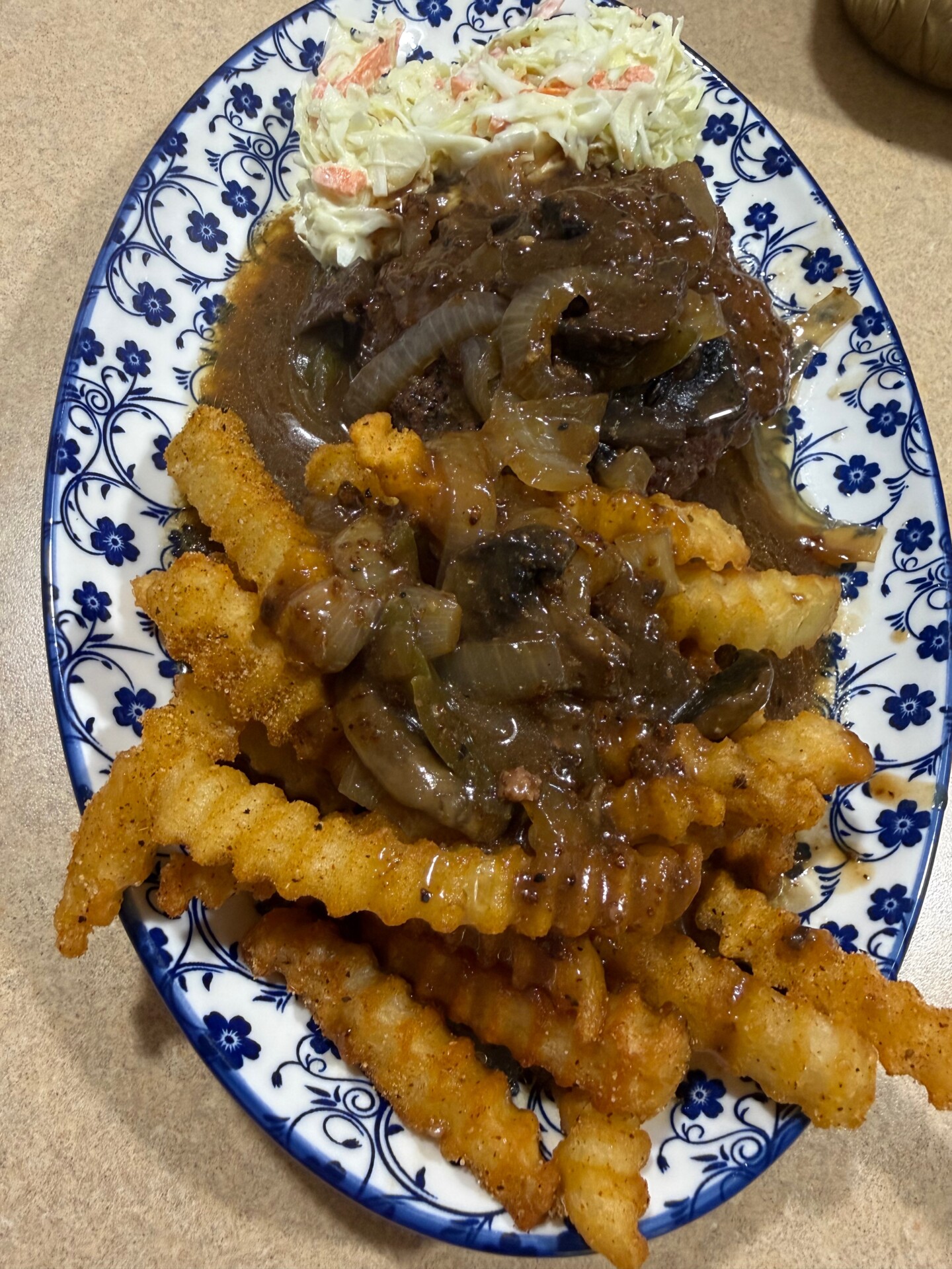 A blue and white patterned plate with crinkle-cut fries, brown gravy with onions and mushrooms over meat, and a serving of coleslaw on the side.