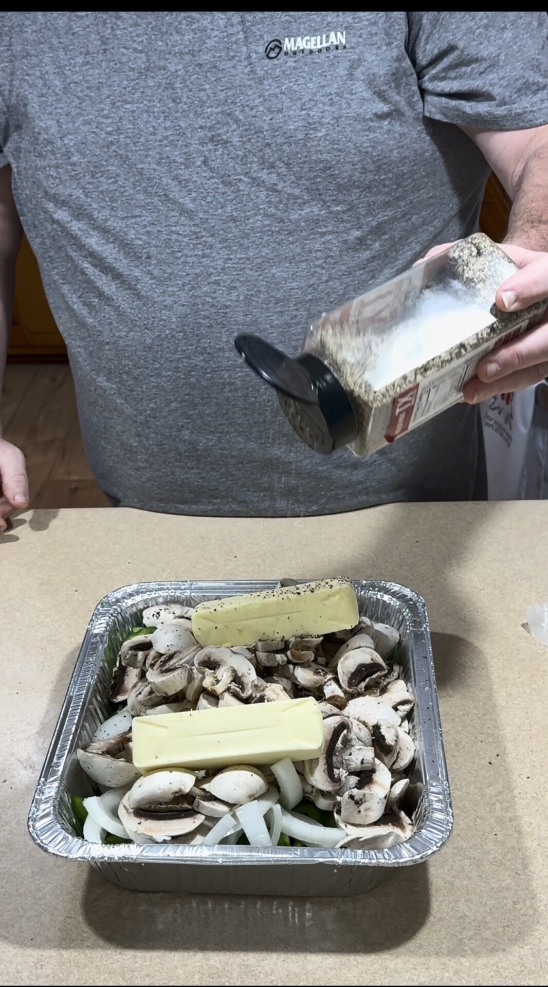 A person in a gray shirt sprinkles seasoning from a container over a foil pan filled with sliced mushrooms, onions, and two sticks of butter on a kitchen counter.