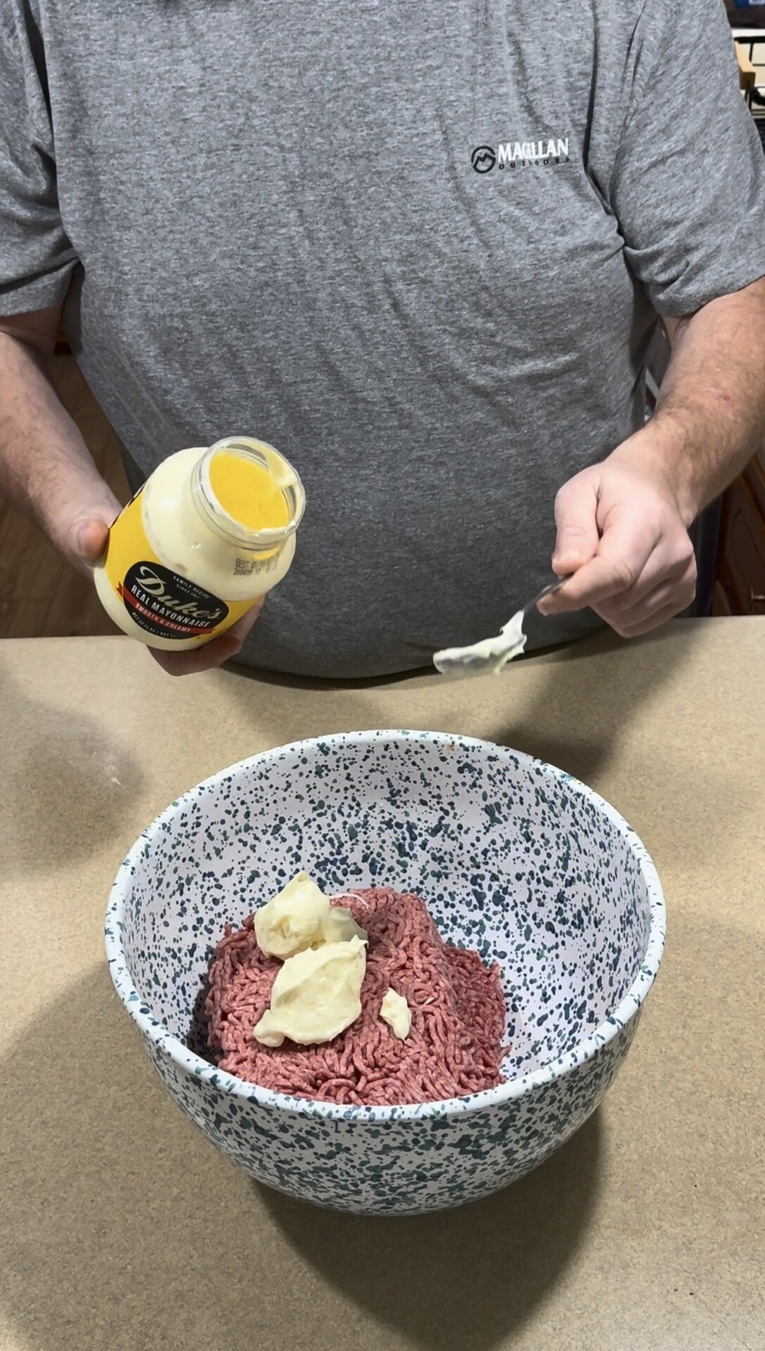 A person in a gray shirt adds a spoonful of mayonnaise from a jar to a bowl containing raw ground beef and more mayonnaise on a kitchen counter.