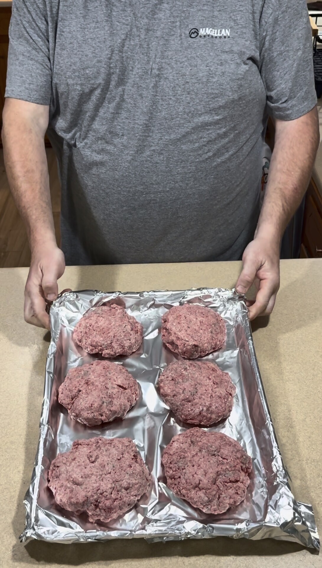 A person in a gray shirt holds a foil-lined baking tray with six raw hamburger patties, evenly spaced, on a kitchen counter.