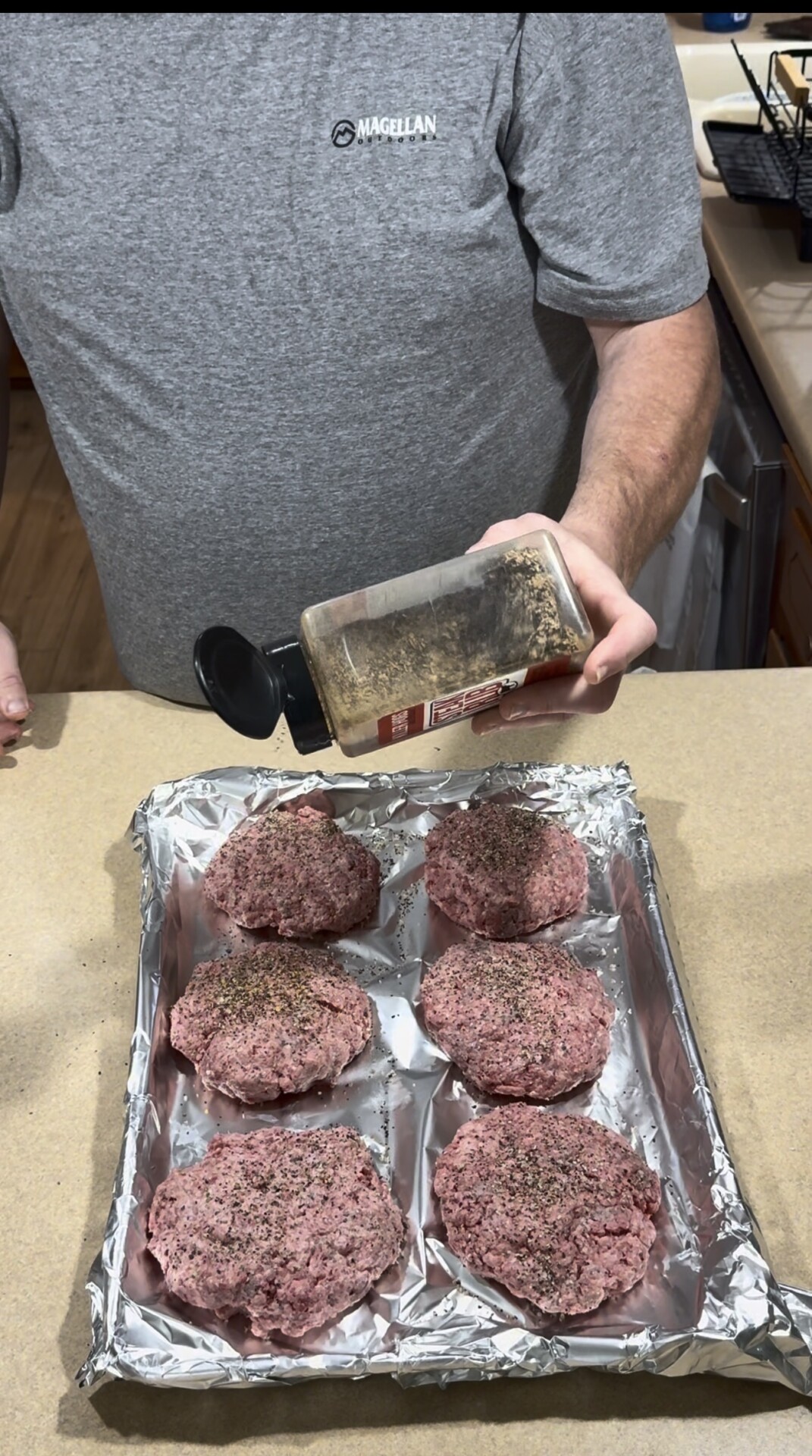 A person sprinkles seasoning onto six raw ground beef patties arranged on a foil-lined baking tray on a kitchen counter.