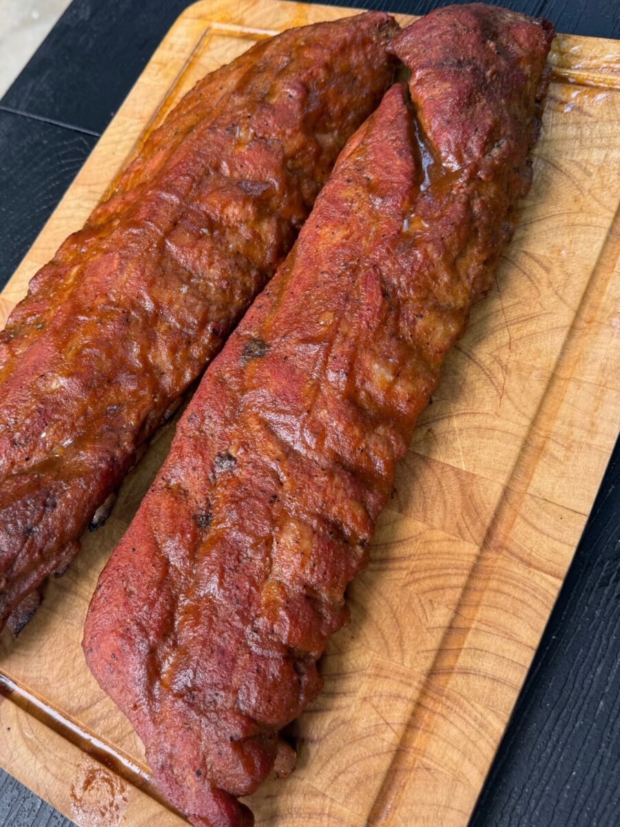 Two racks of smoked barbecue ribs with a reddish glaze rest on a wooden cutting board, placed on a dark surface. The ribs appear juicy and well-cooked, with visible grill marks and seasoning.
