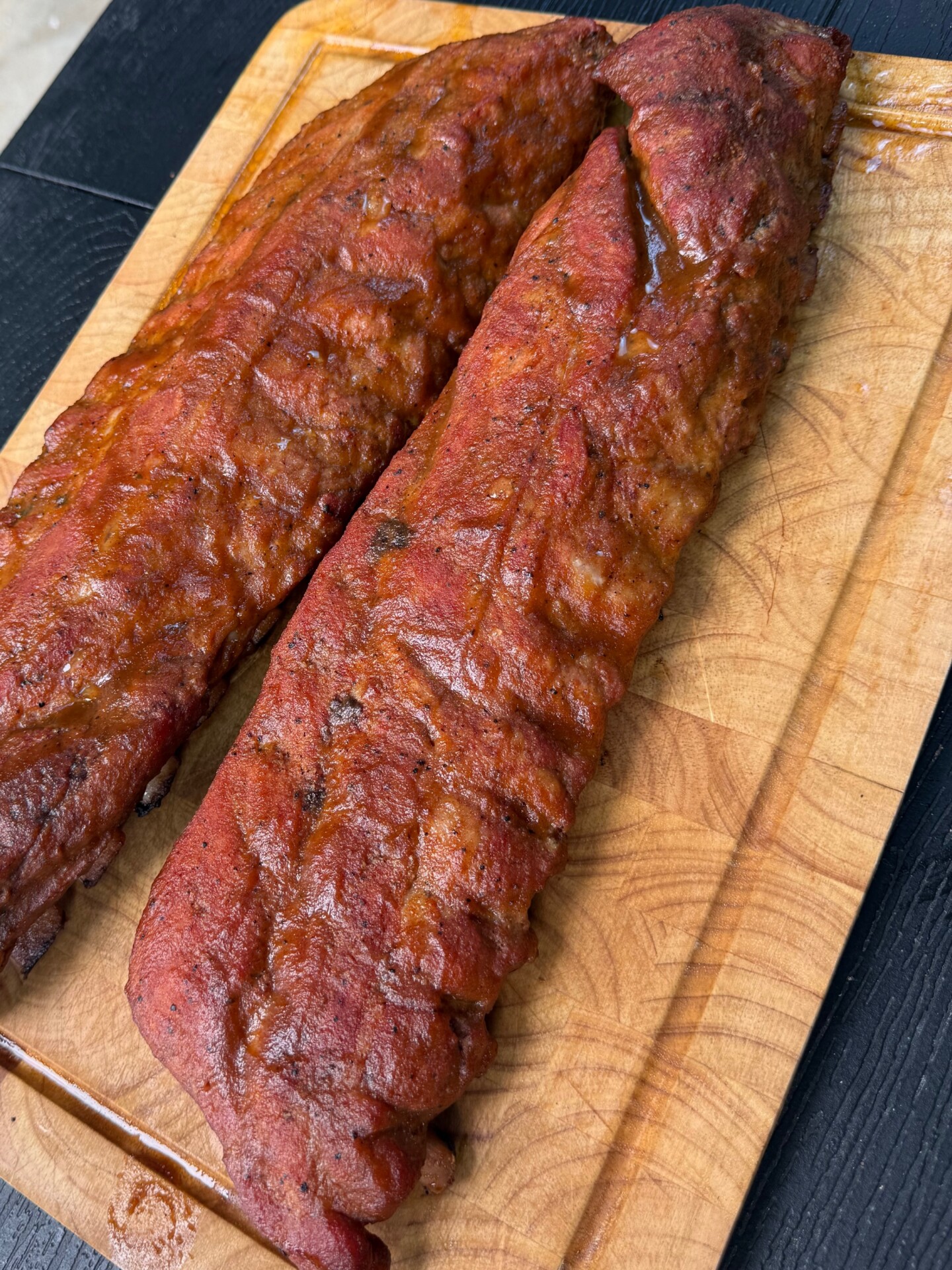 Two racks of smoked barbecue ribs with a reddish glaze rest on a wooden cutting board, placed on a dark surface. The ribs appear juicy and well-cooked, with visible grill marks and seasoning.