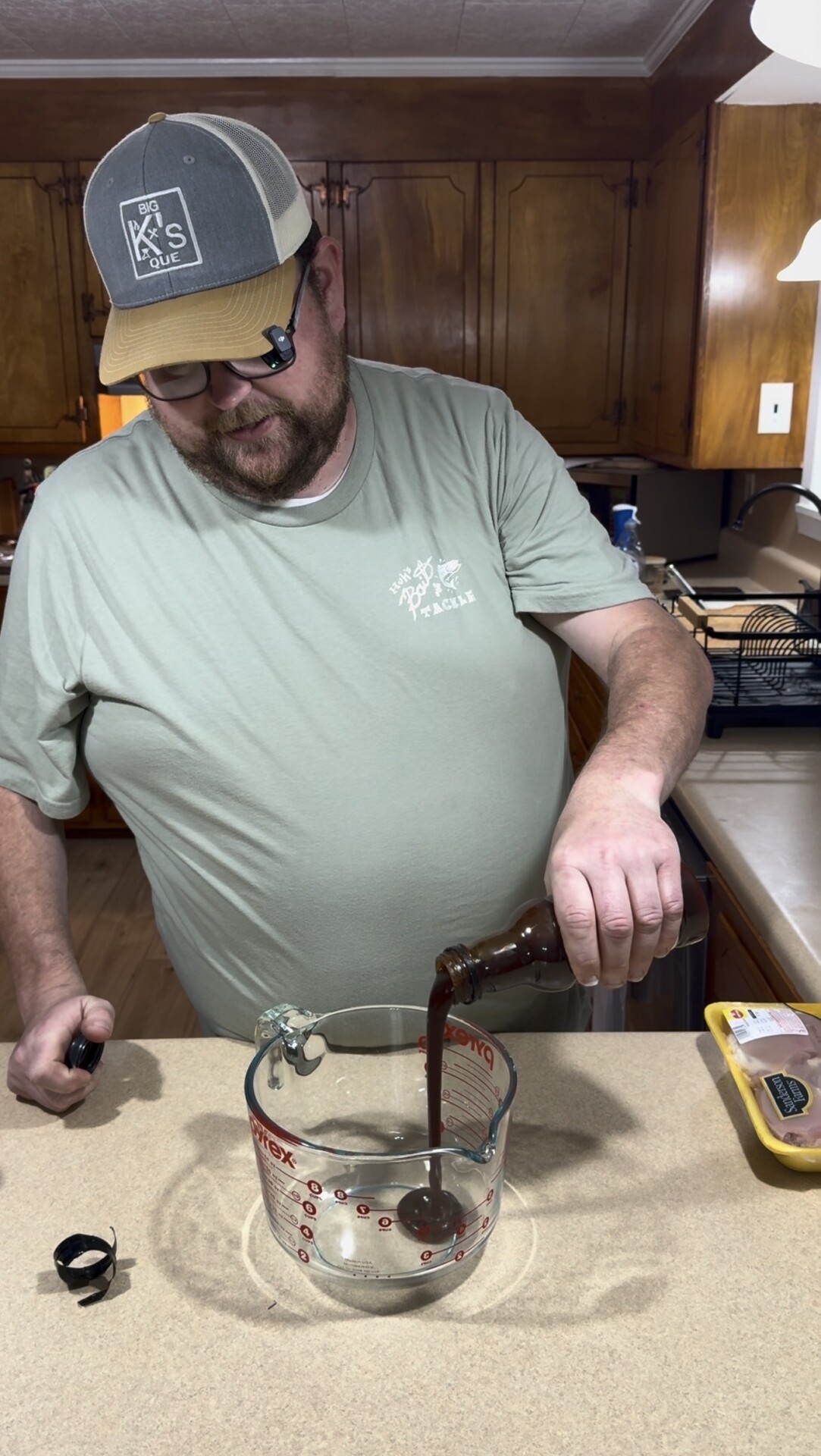 A man wearing a green T-shirt and a cap pours a dark liquid from a bottle into a glass measuring cup on a kitchen counter, with a pack of raw chicken nearby.