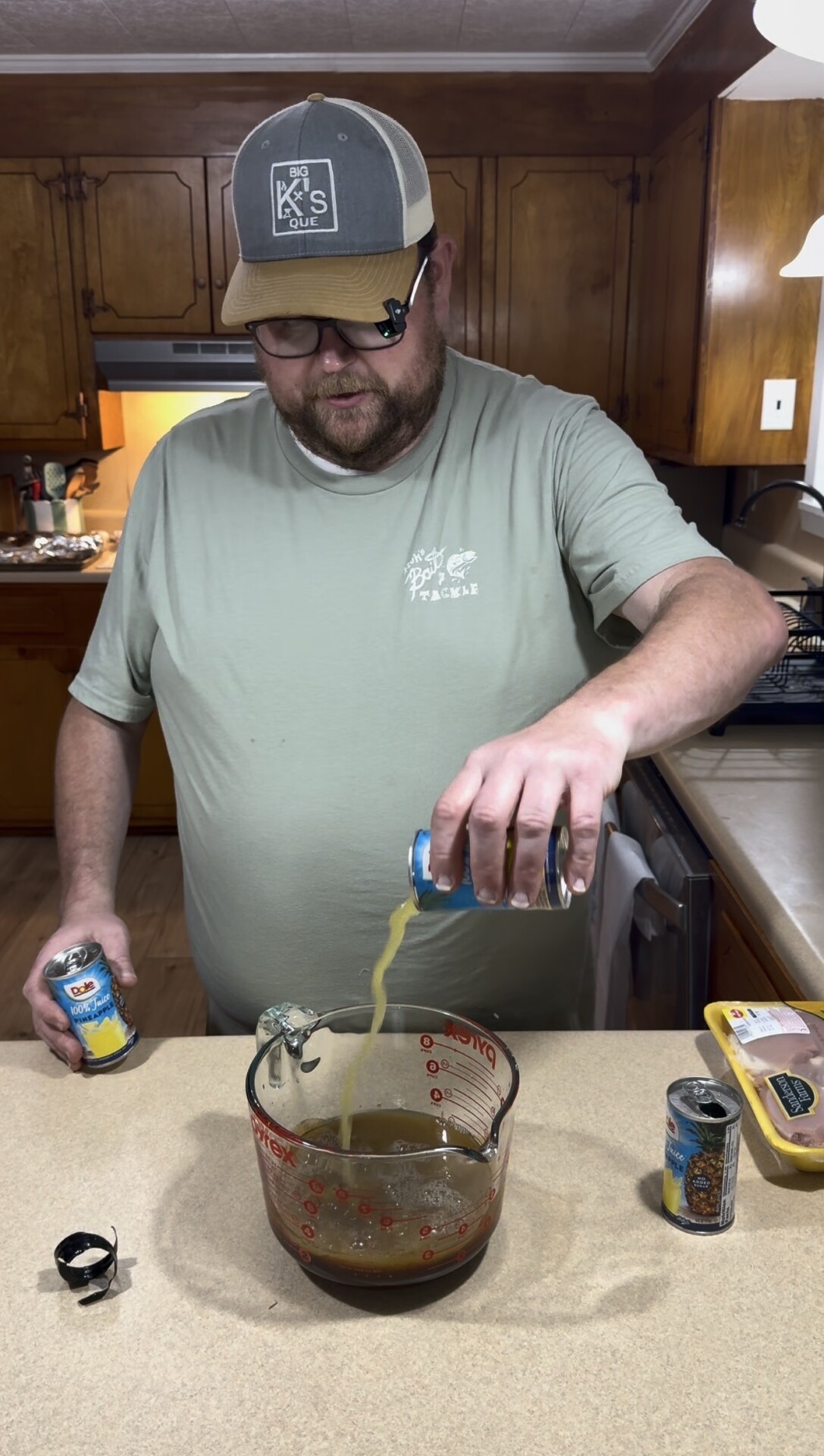 A man wearing a cap and glasses stands in a kitchen, pouring liquid from a can into a large measuring cup on the counter. Other cans and a package of chicken are nearby.