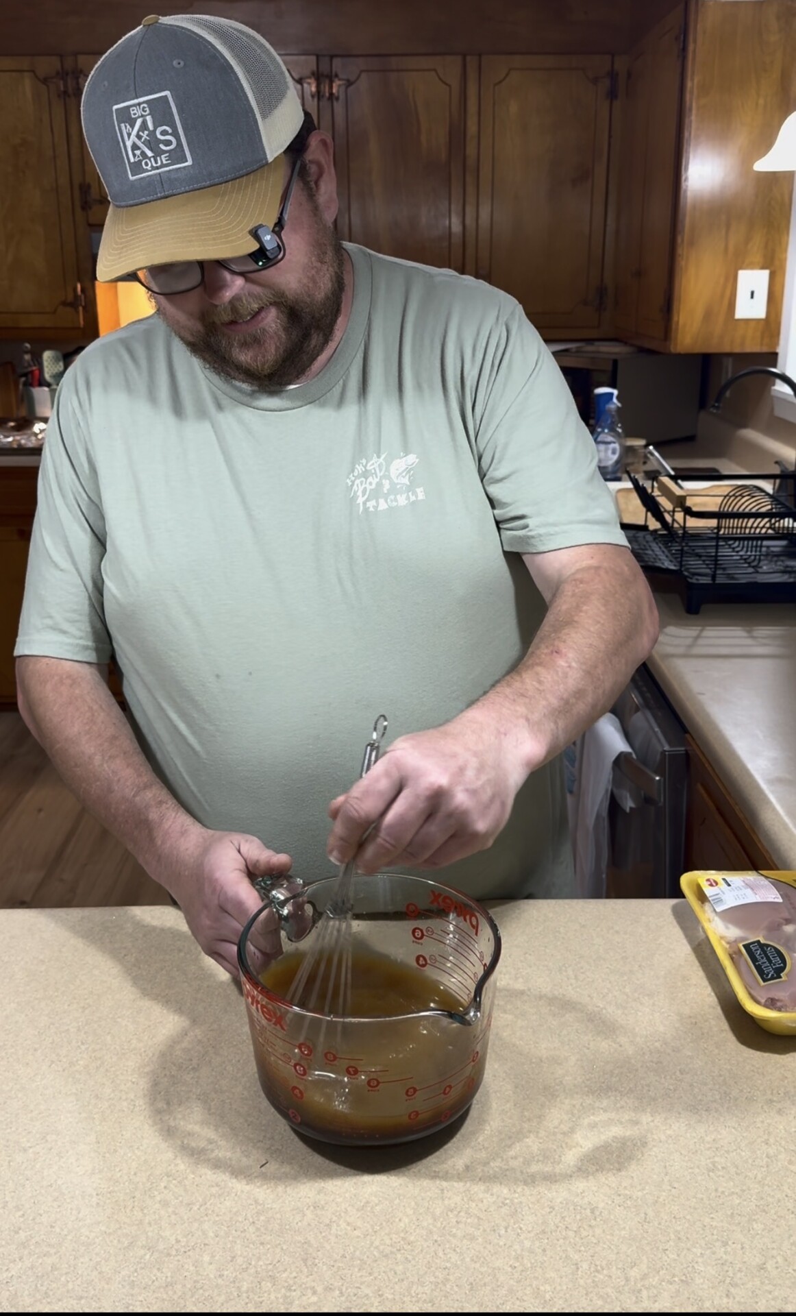 A man in a green T-shirt and cap is using a whisk to mix a brown liquid in a large glass measuring cup on a kitchen counter. A package of raw chicken is on the counter nearby.
