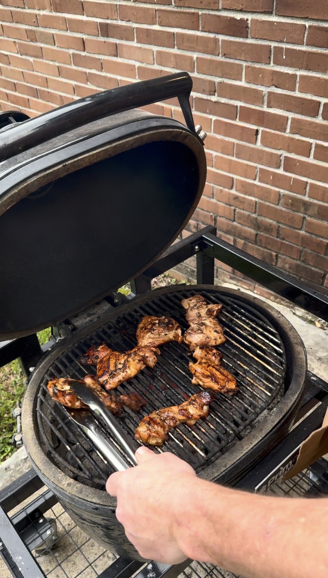 A person grills marinated meat on an open charcoal barbecue grill, using tongs to turn the pieces. The grill is next to a brick wall and set outdoors.