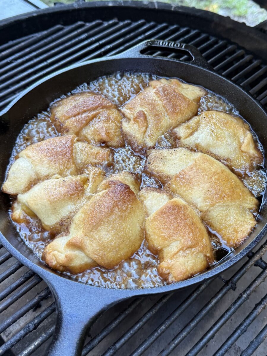 Golden brown crescent rolls are baking in a cast iron skillet, bubbling in a caramelized sauce, set on a grill outdoors.
