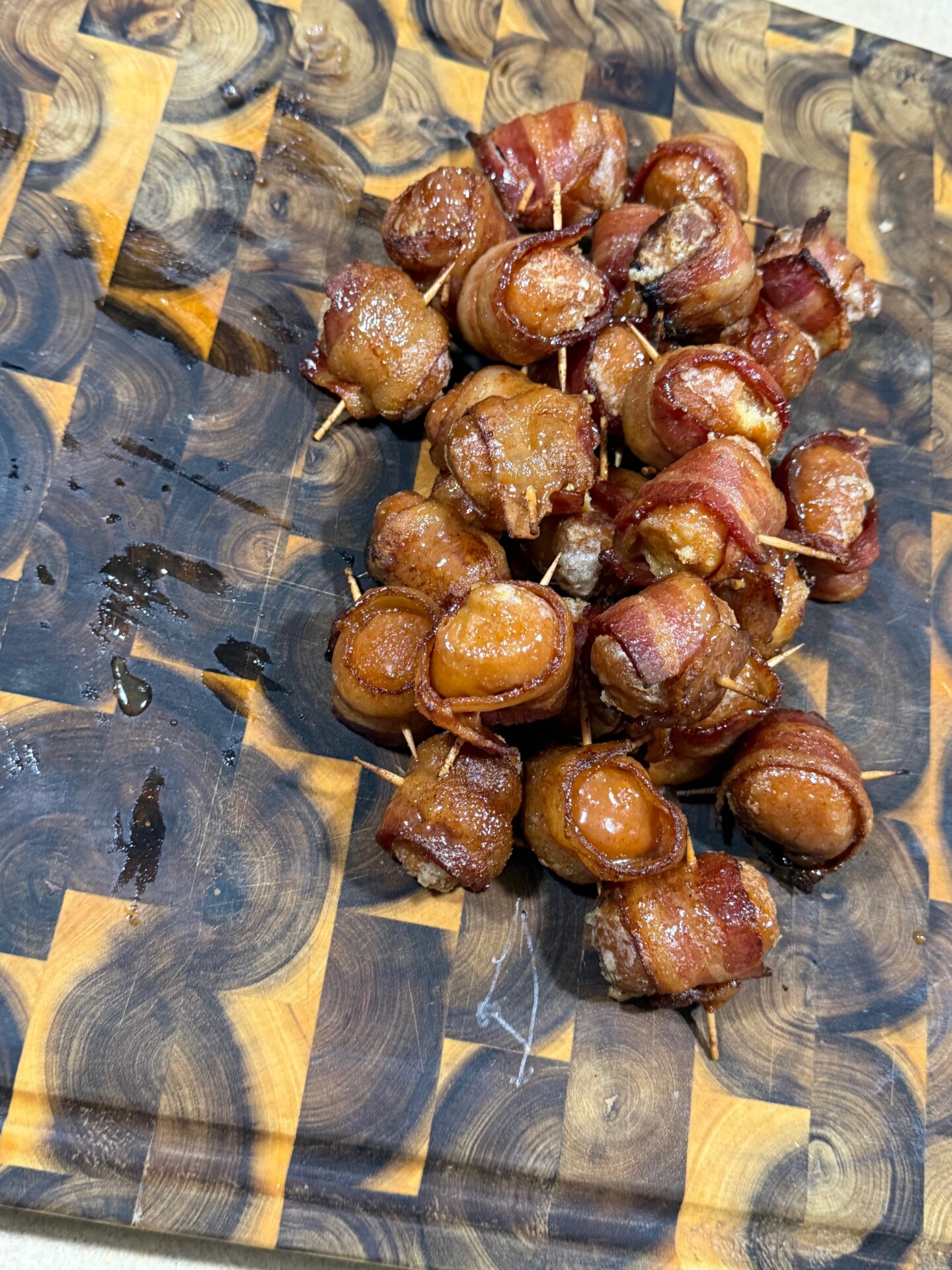 A pile of bite-sized sausages wrapped in crispy bacon, secured with toothpicks, sits on a patterned wooden cutting board. The bacon appears caramelized and golden brown.