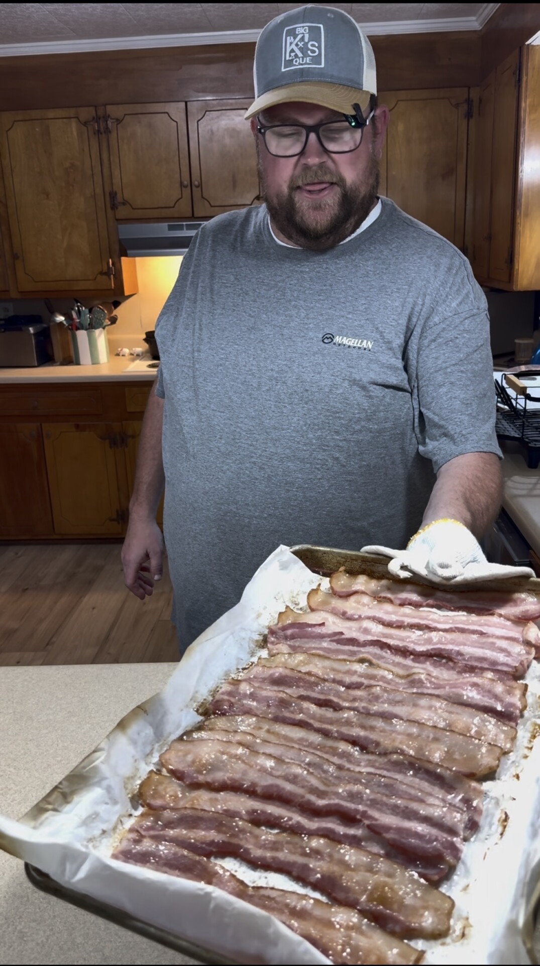 A man wearing a gray shirt and hat smiles in a kitchen, holding a baking sheet covered with strips of cooked bacon on parchment paper. Wooden cabinets and kitchen utensils are visible in the background.