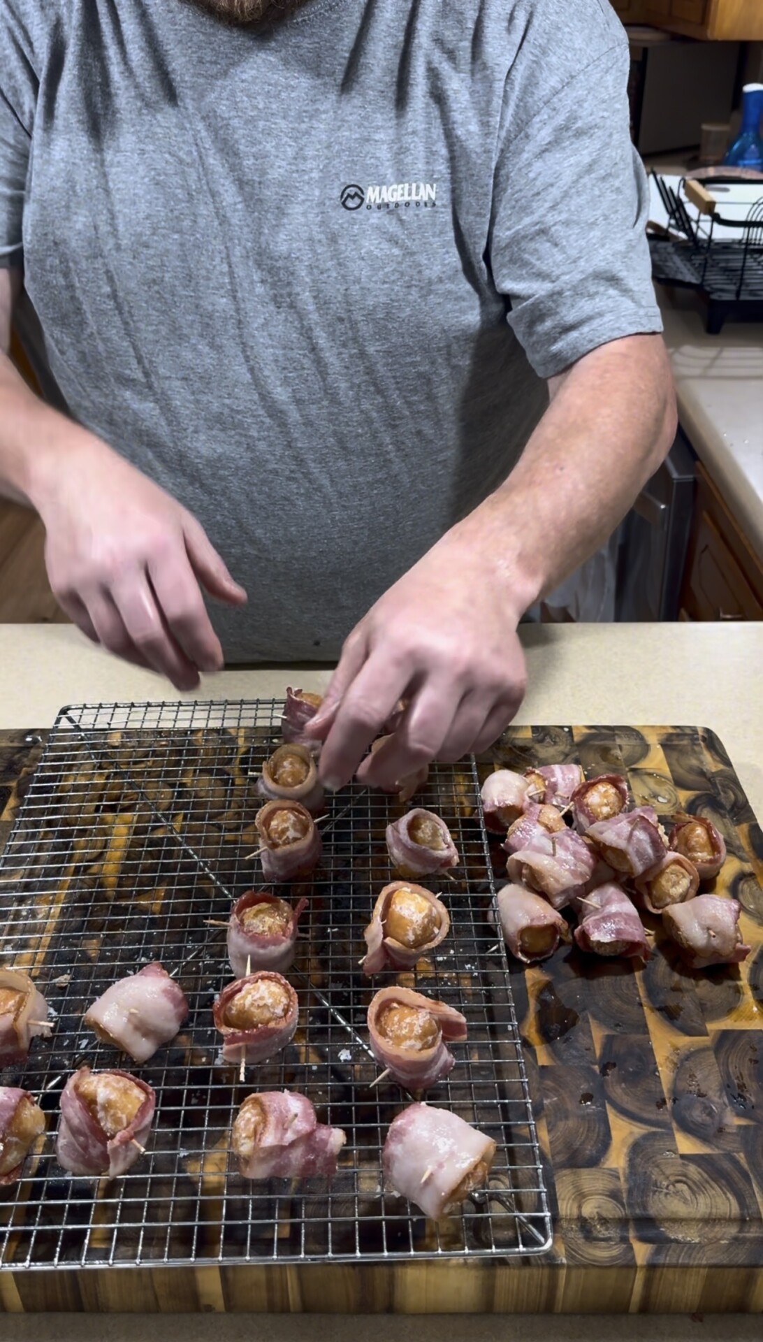 A person wearing a gray shirt places bacon-wrapped food pieces onto a wire rack set over a baking sheet in a kitchen. More bacon-wrapped pieces are on the cutting board nearby.