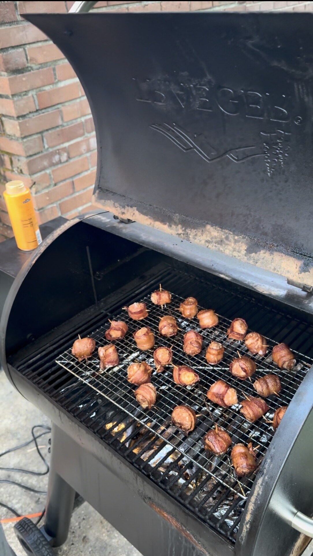 A smoker grill with its lid open, showing several bacon-wrapped bites on a wire rack being cooked. A can of seasoning or spray sits on the grill’s side shelf, and theres a brick wall in the background.