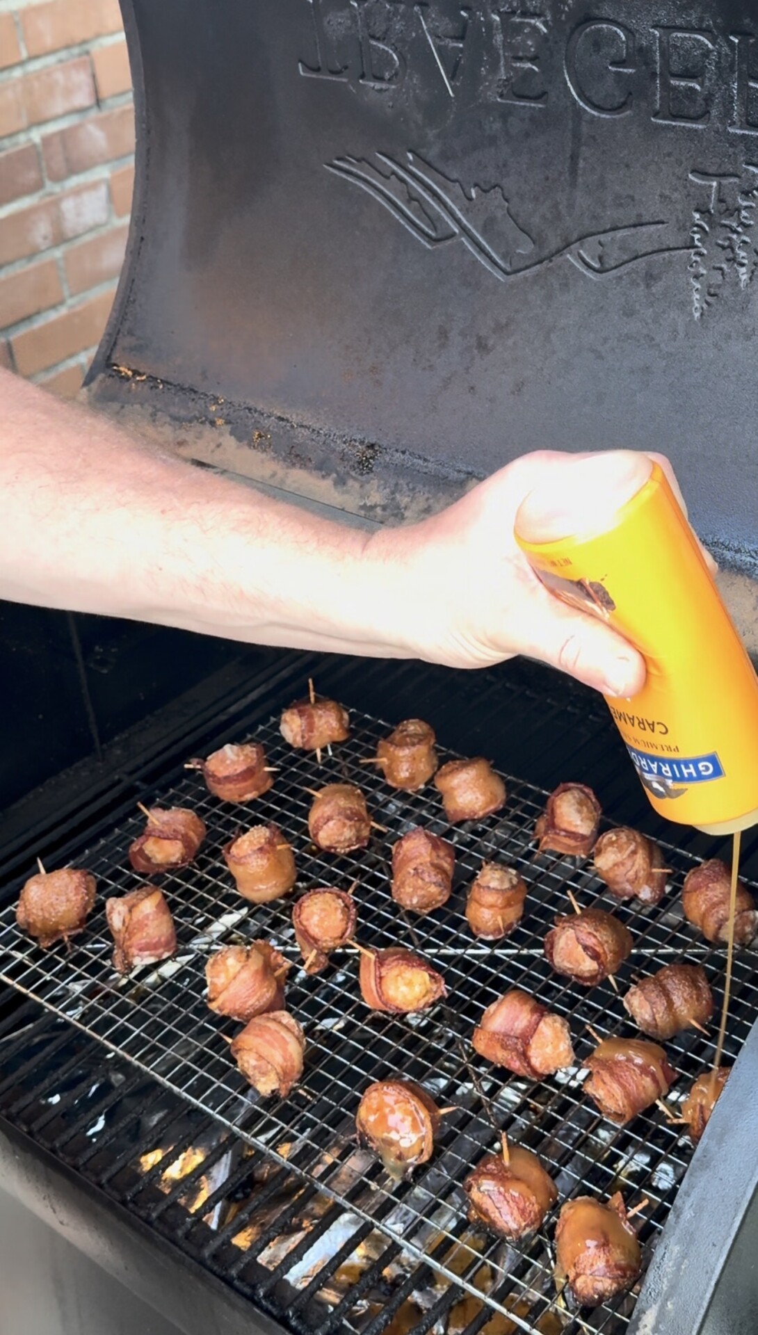 A hand pours a can of beer over bacon-wrapped food items on a grill, with the grill lid open and food cooking on wire racks. Brick wall is visible in the background.