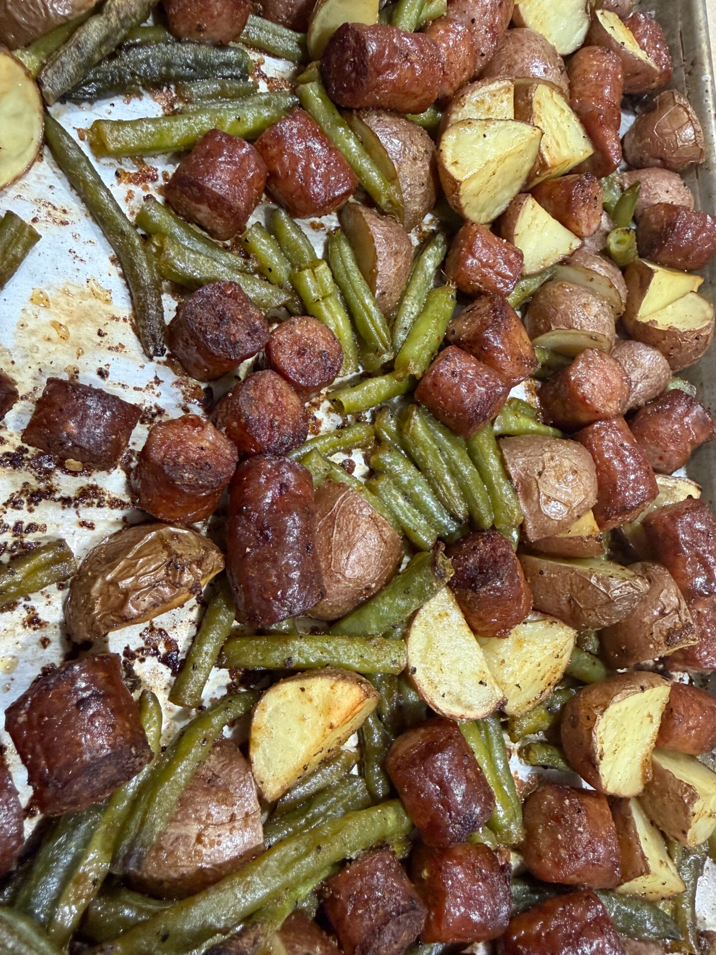 A sheet pan filled with roasted green beans, sliced sausage, and quartered red potatoes, all browned and cooked together.