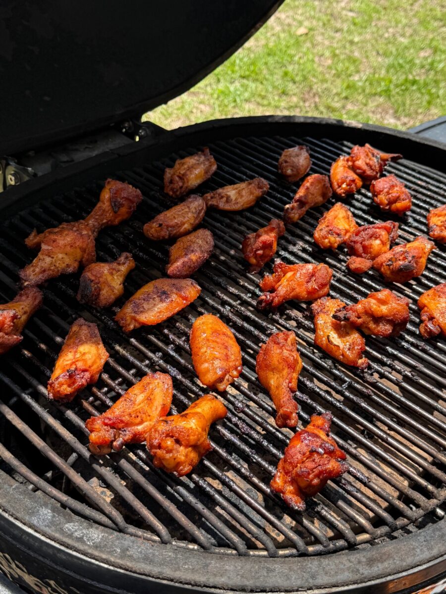 Chicken wings and drumsticks coated in seasoning are grilling on an open barbecue. The meat is arranged across the grates, cooking over indirect heat, with a lawn visible in the background.