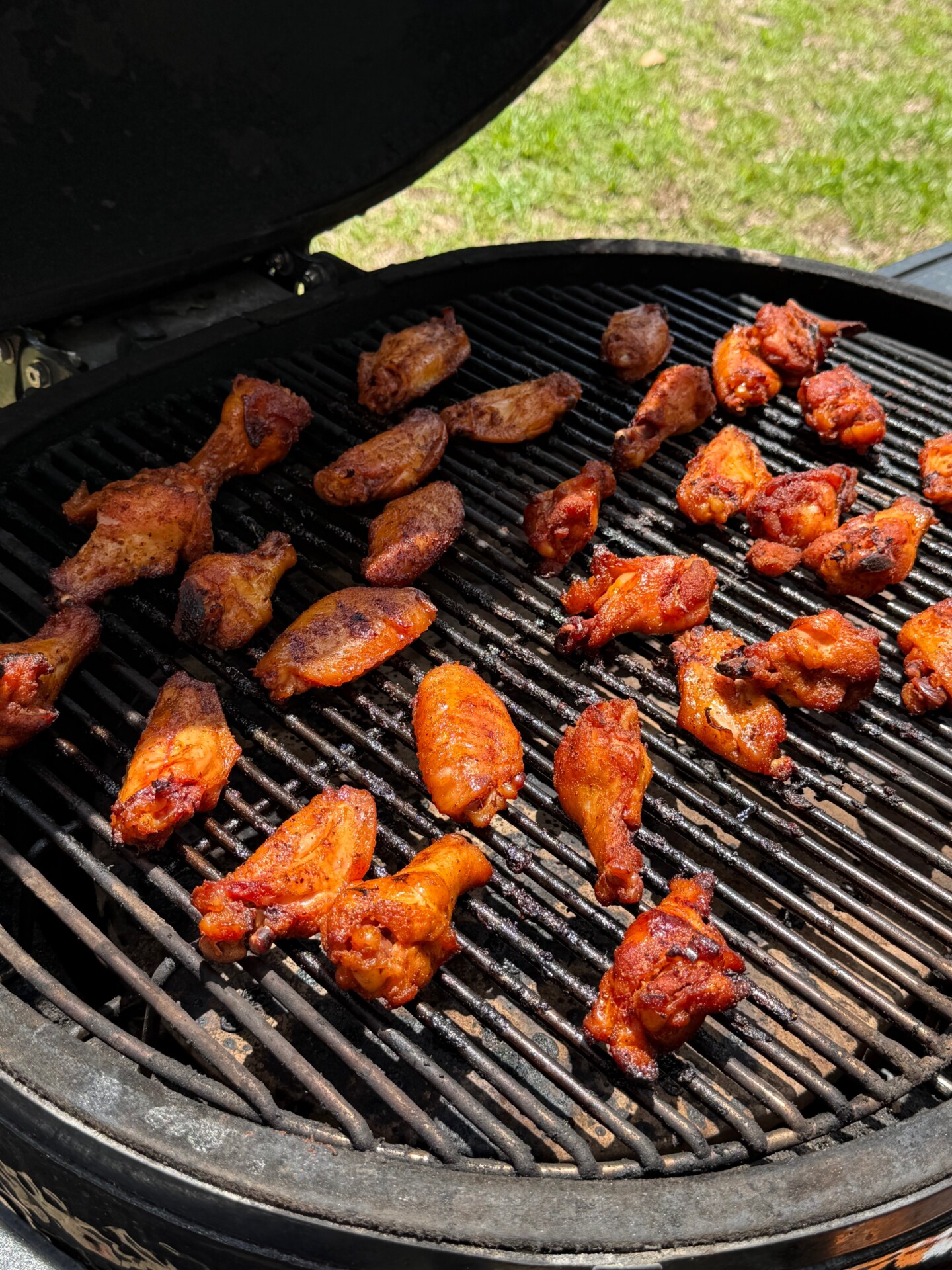 Chicken wings cooking on a charcoal grill with a dark lid open, grill marks visible on the wings, and a green grassy area in the background.