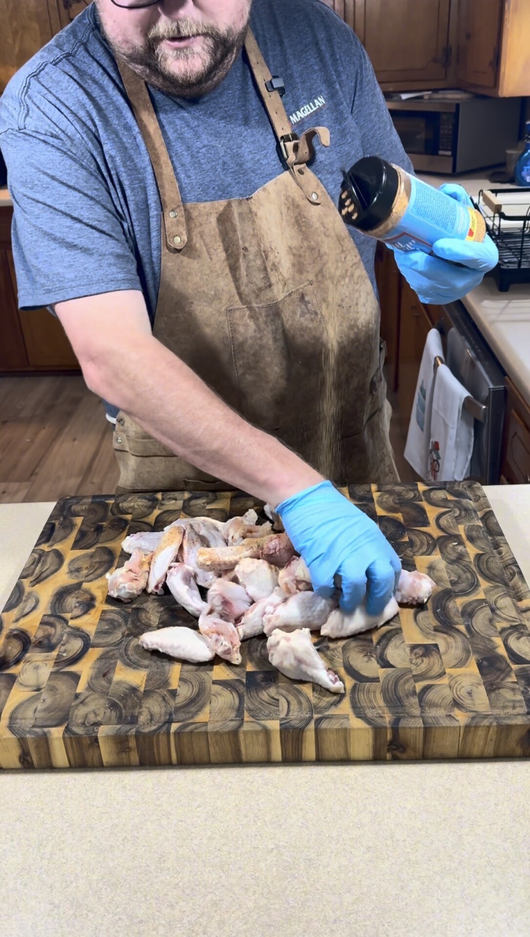 A person wearing a brown apron and blue gloves seasons raw chicken wings on a patterned wooden cutting board in a kitchen.