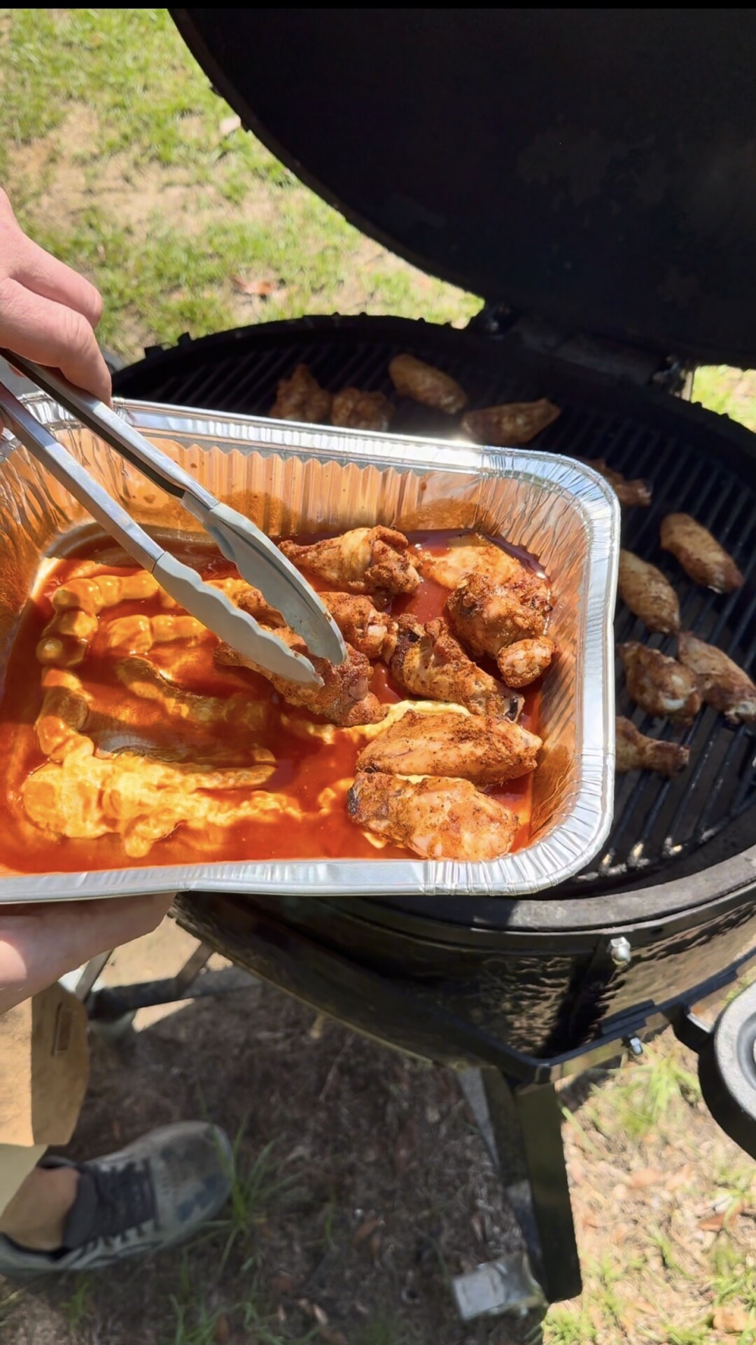 A person uses tongs to add grilled chicken wings to a foil tray filled with orange sauce, next to an open charcoal grill with more wings cooking. The scene is outdoors in daylight.