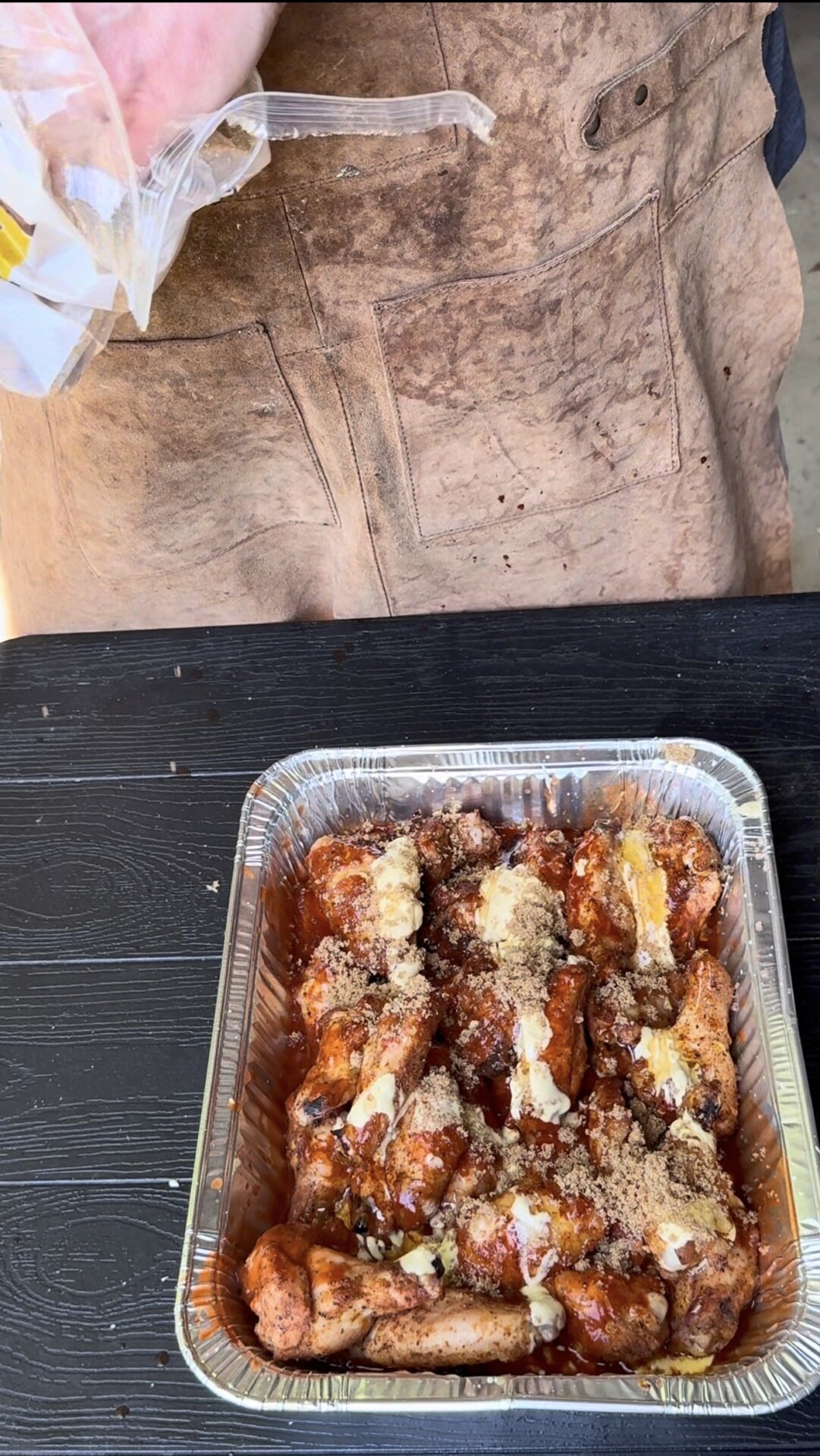 A person wearing a brown apron is sprinkling ingredients onto raw, seasoned chicken drumsticks in a disposable foil tray on a black table. The chicken is covered with spices, brown sugar, and butter pieces.