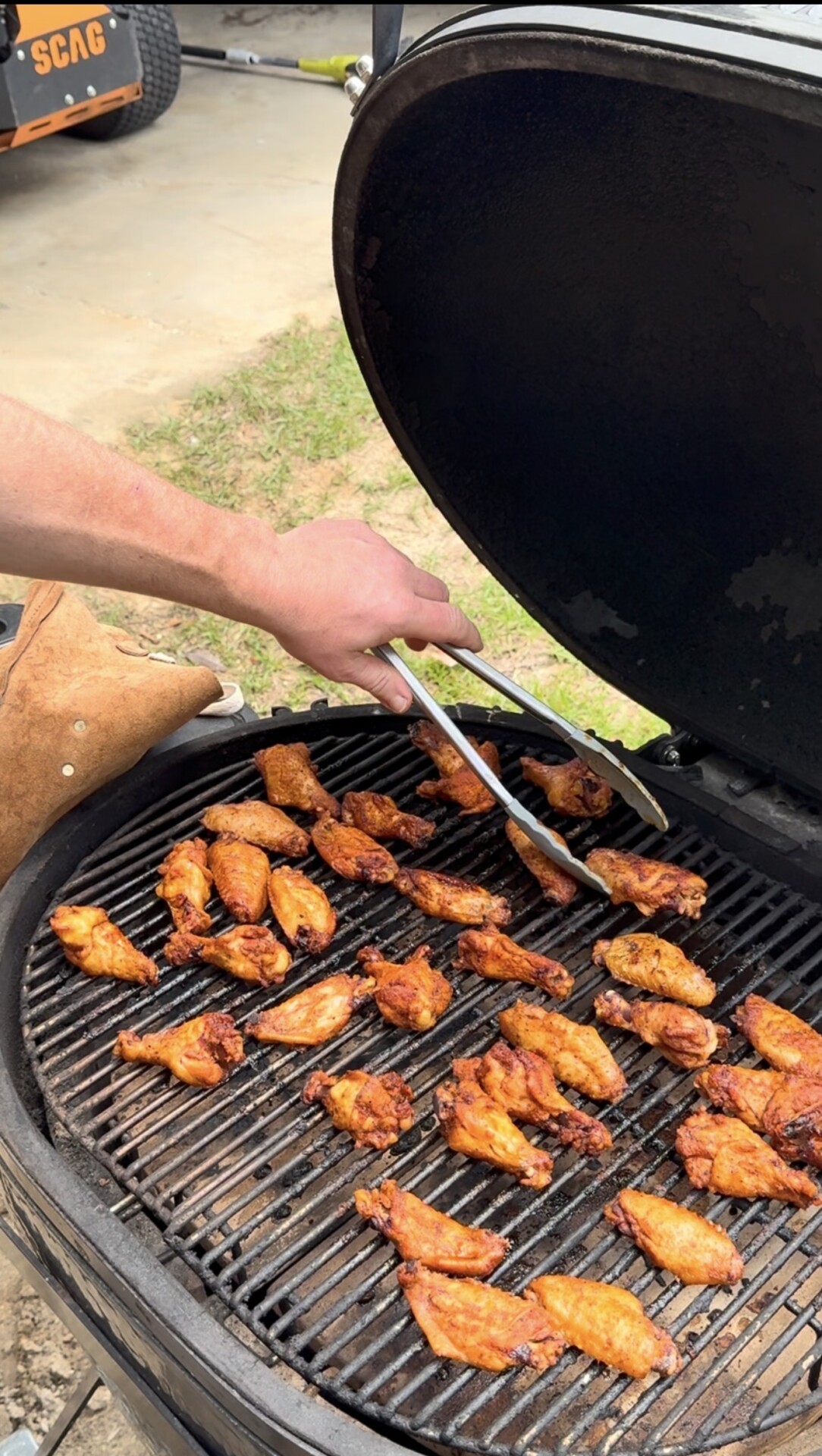 A person uses tongs to grill several seasoned chicken wings on a round barbecue grill with the lid open, outdoors on a grassy area.