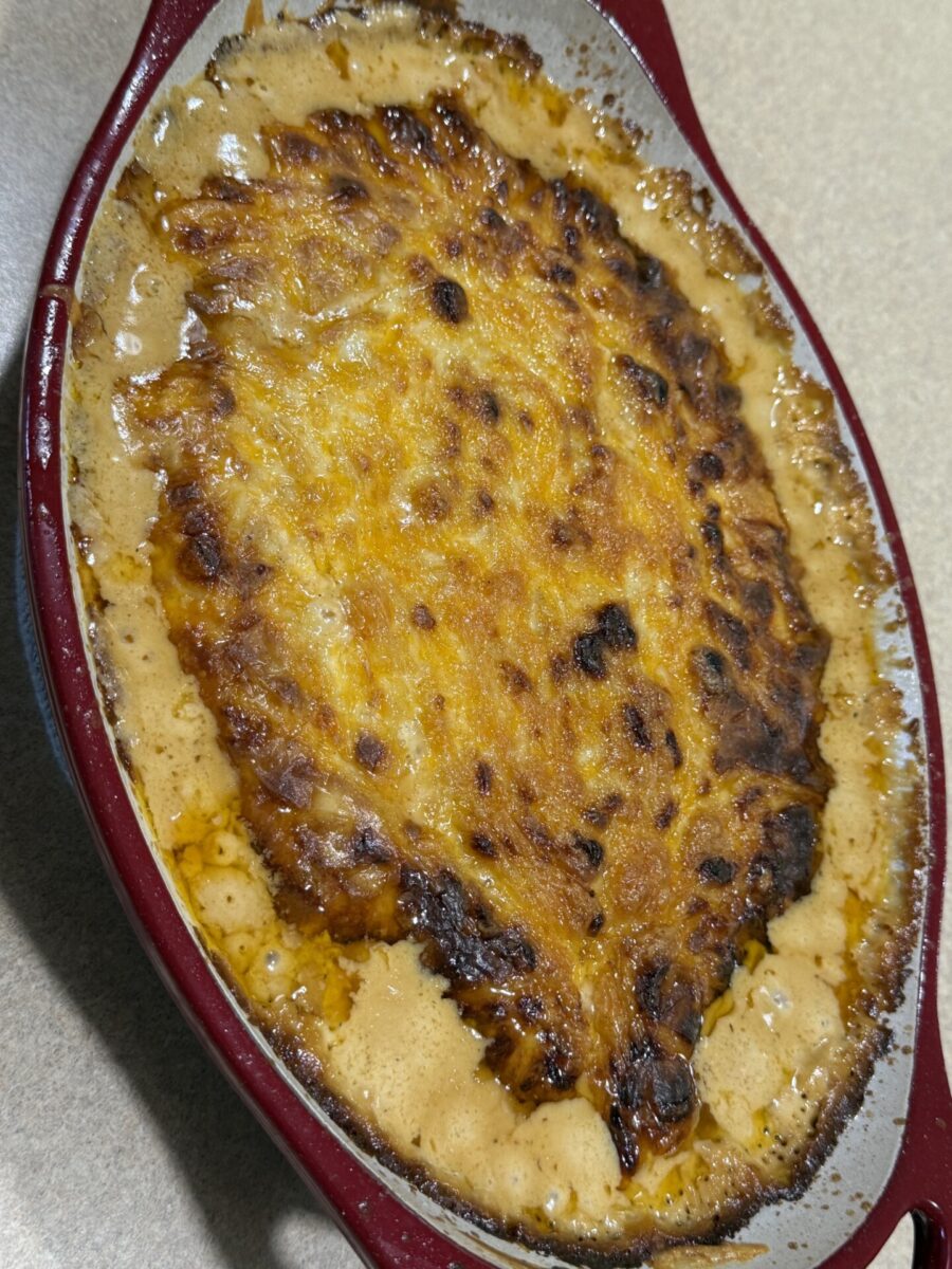 A baked casserole with a golden brown, slightly crispy top sits in an oval red dish. The edges show bubbling, melted cheese and sauce. The dish rests on a beige countertop.