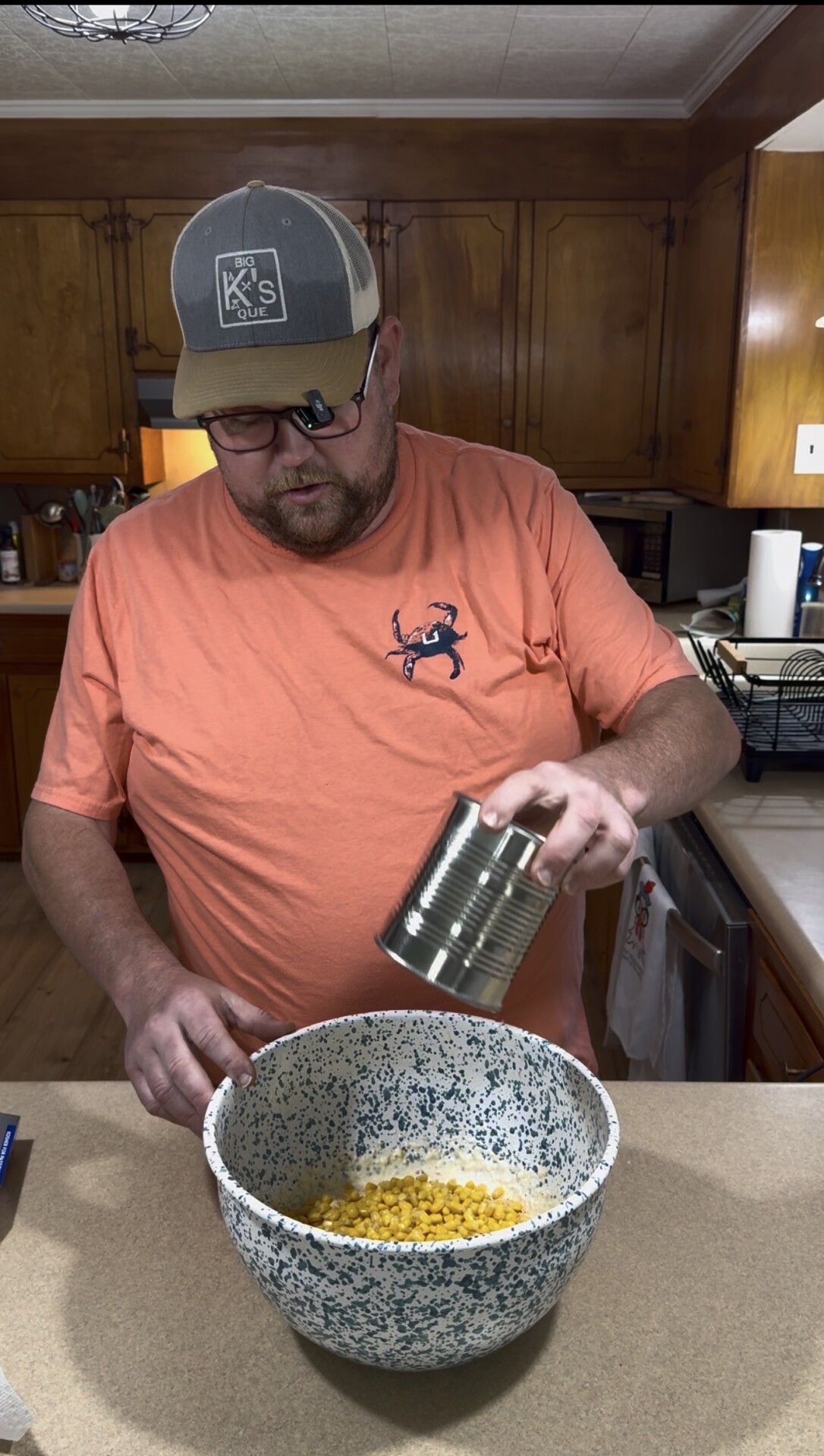 A man in an orange shirt and cap pours canned corn into a large mixing bowl on a kitchen counter, with wooden cabinets and dishes visible in the background.